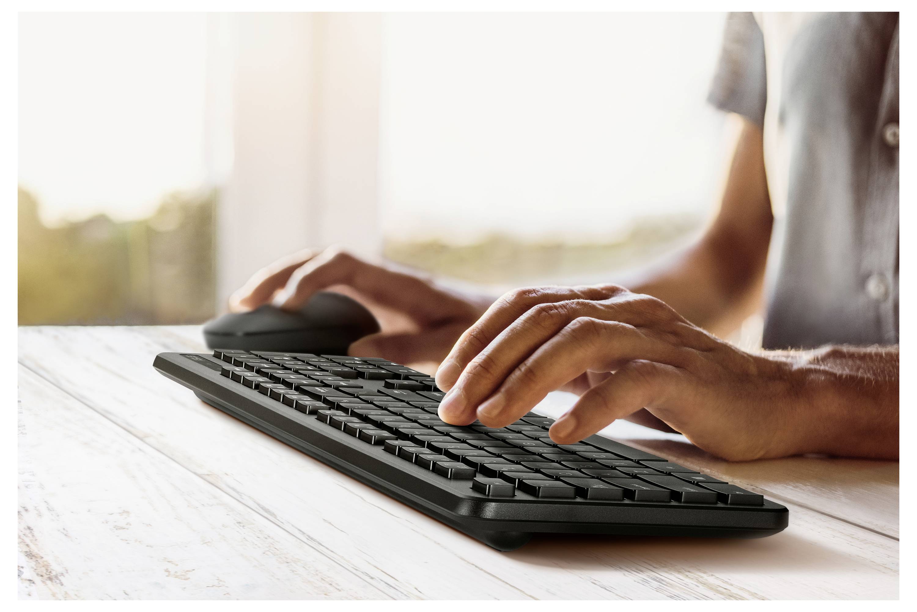 Osoba pisząca na bezprzewodowej klawiaturze z myszką w pobliżu, w jasnym pokoju z widokiem na okno.