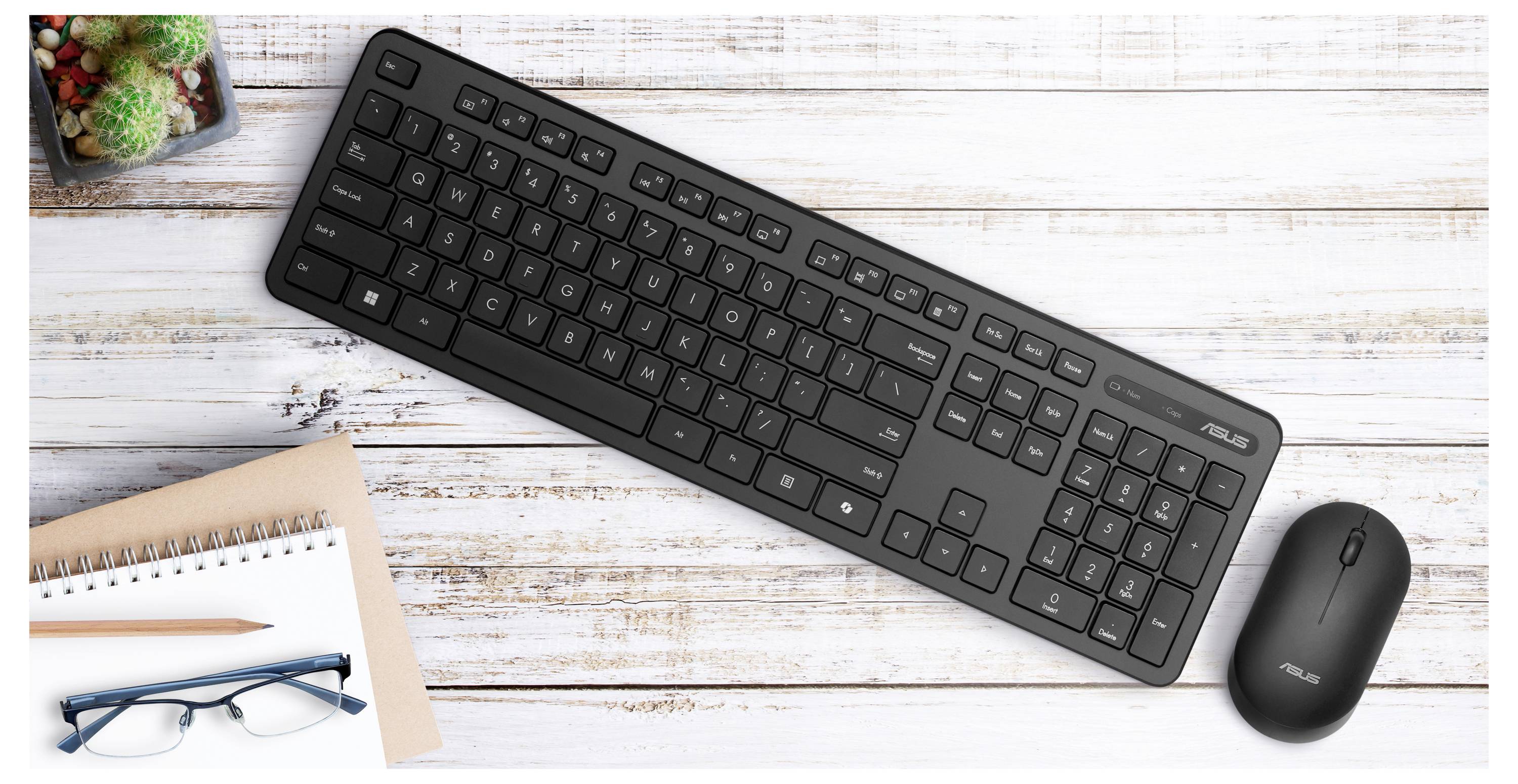 Czarny zestaw klawiatury i myszy umieszczony na białym drewnianym stole, obok notesu, długopisu, okularów i małych roślin.