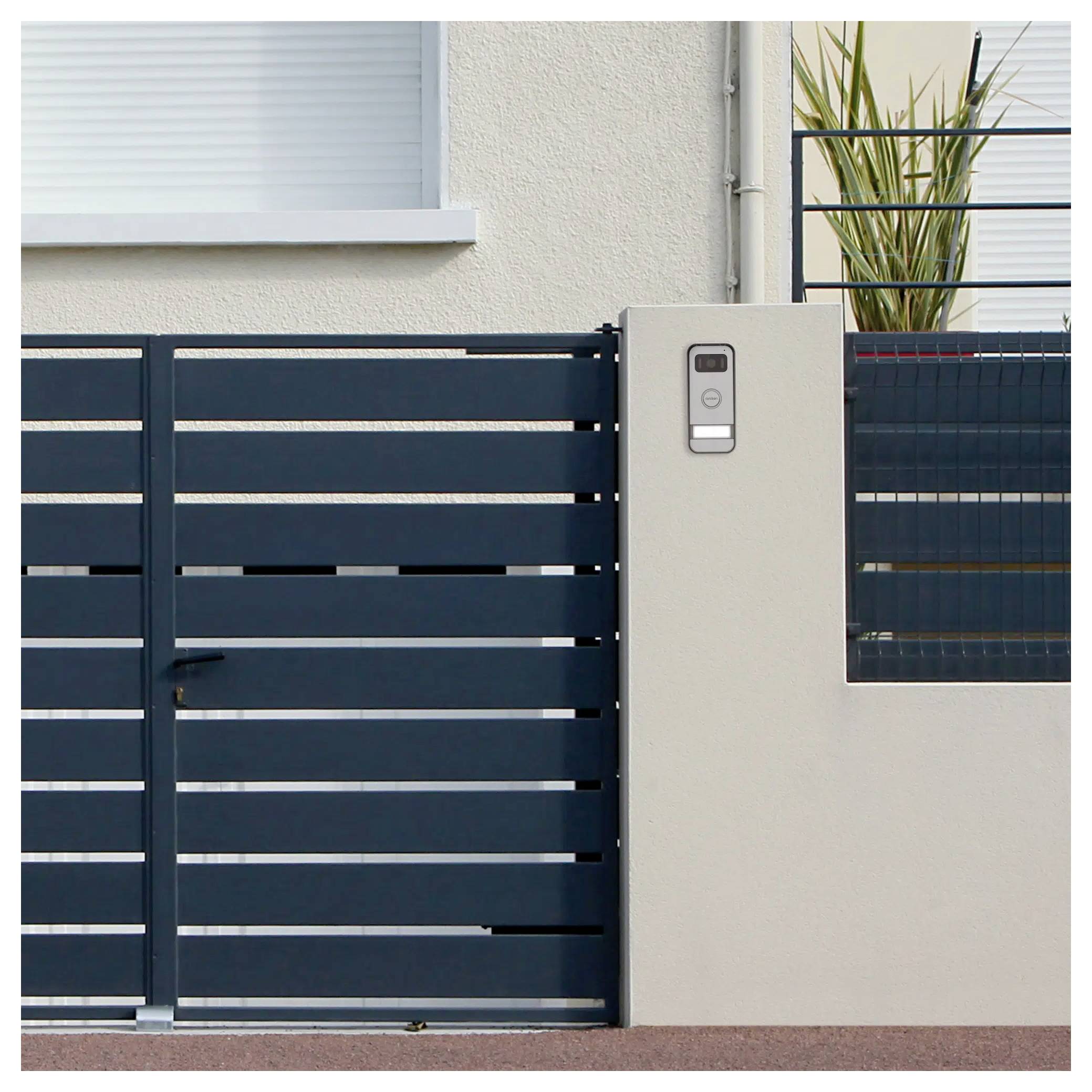 Nowoczesna czarna metalowa brama i domofon na betonowym słupie przed białym budynkiem, z doniczkową rośliną na balkonie powyżej.