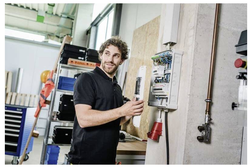 Technik sprawdzający panel sterowania elektrycznego w warsztacie, trzymający otwarte drzwi w pobliżu narzędzi i półek do przechowywania.