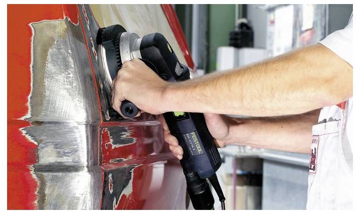 Osoba używająca szlifierki elektrycznej na czerwonej powierzchni metalowej w warsztacie, wygładzająca krawędzie w celu uzyskania wykończenia na wysokim poziomie.