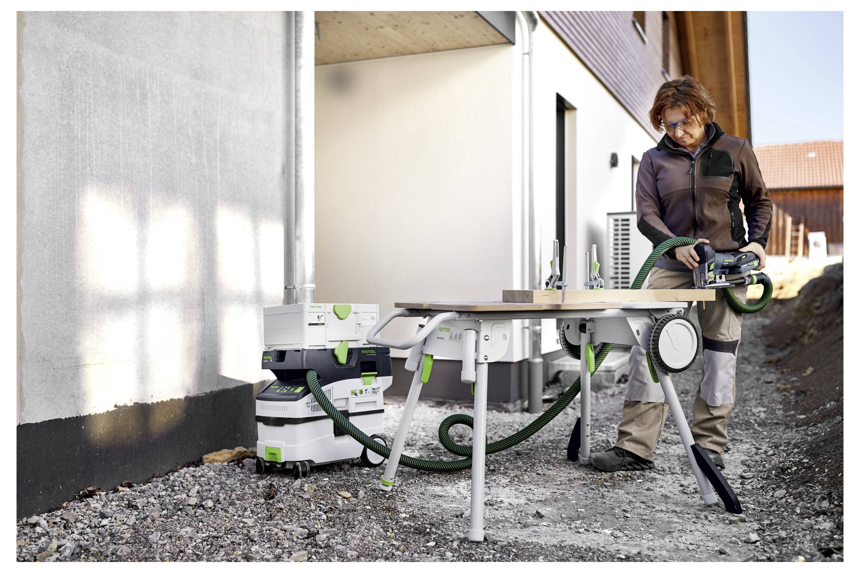Osoba pracuje z narzędziem elektrycznym podłączonym do systemu odciągającego pył na improwizowanym stole roboczym na zewnątrz, w pobliżu budynku będącego w trakcie budowy.