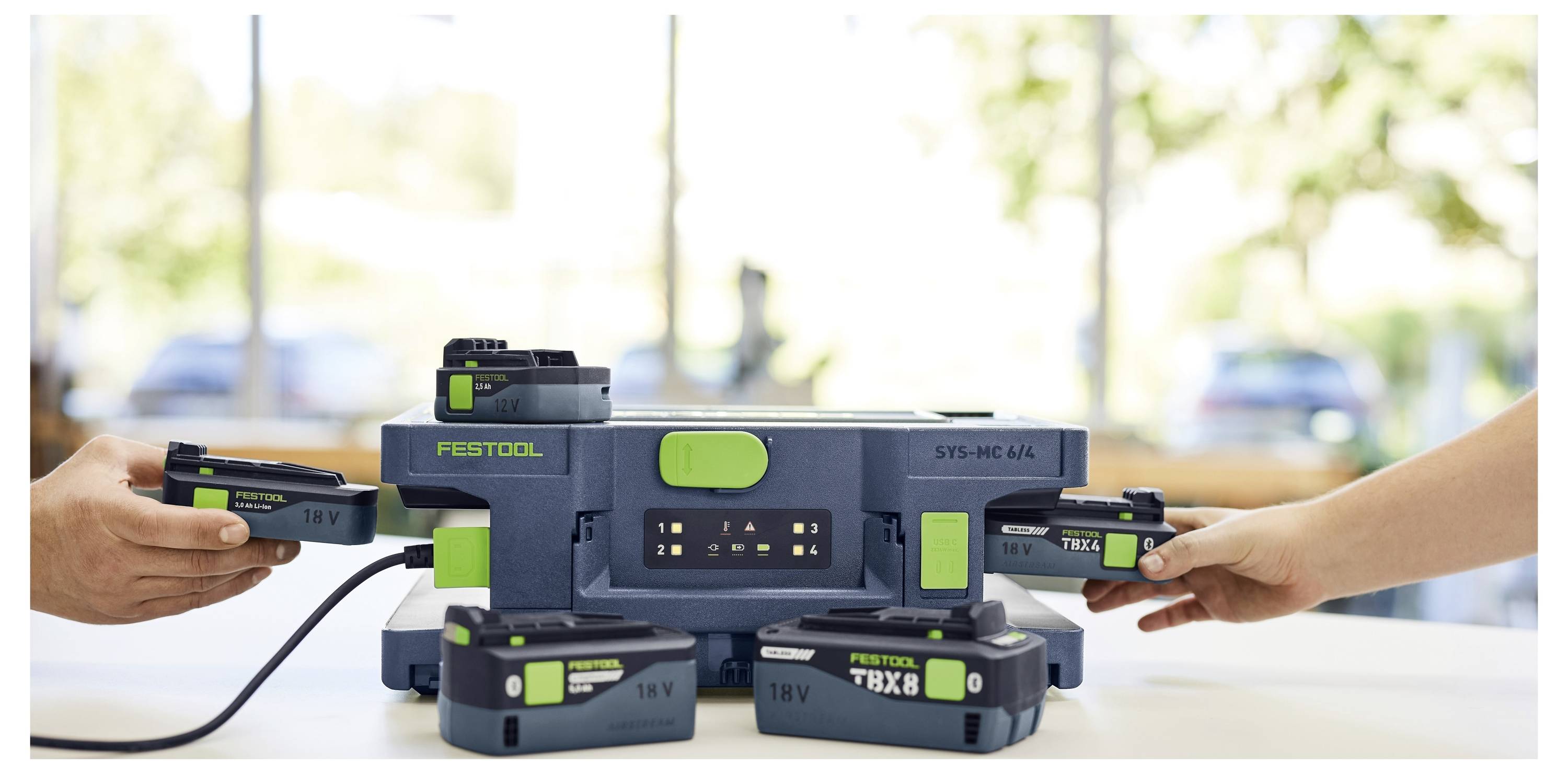 Dwie ręce wkładają baterie do Festool SYS-PowerStation. Stacja pokazuje wiele ładowań w jasnym, nasłonecznionym pomieszczeniu.