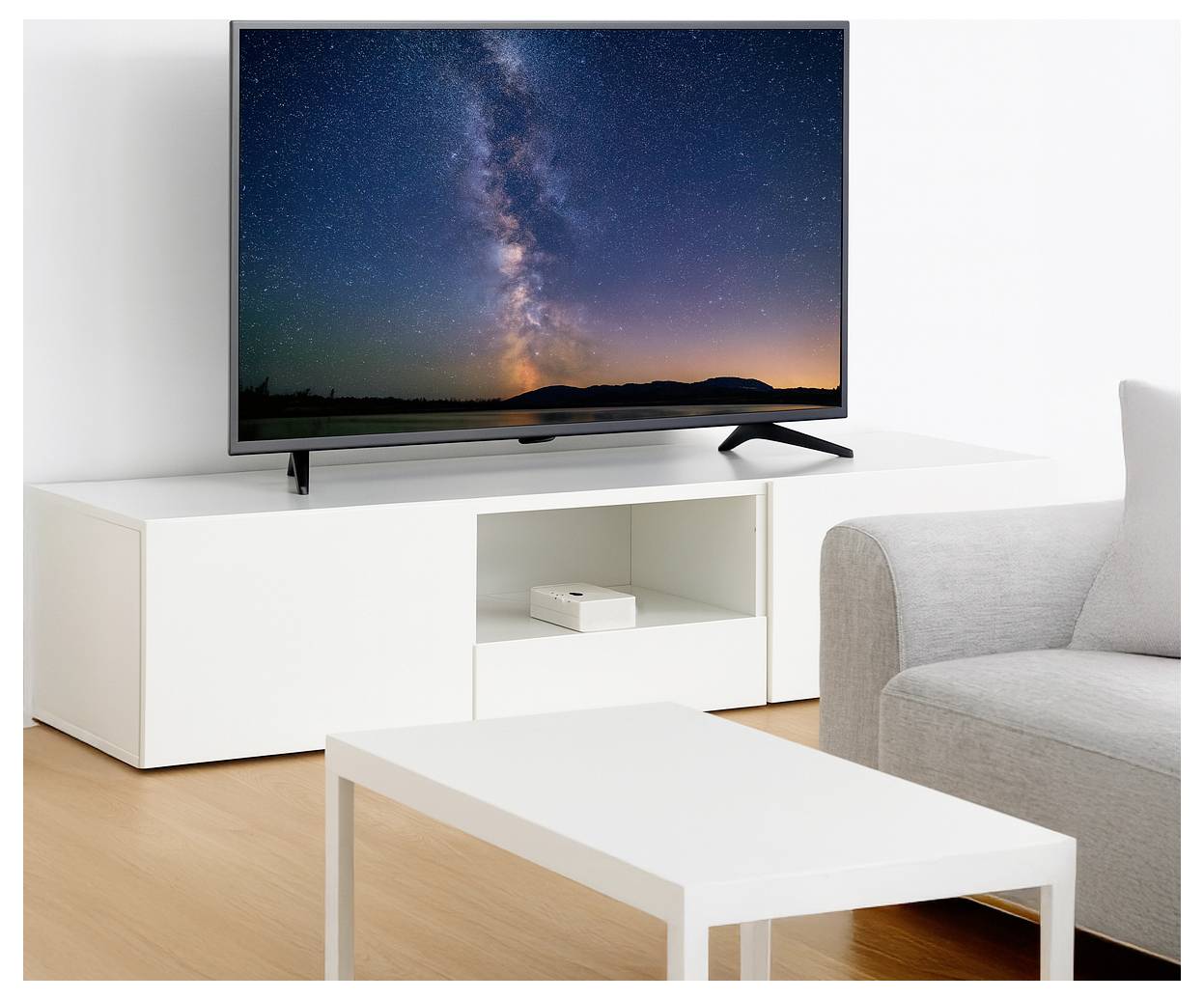 Telewizor z płaskim ekranem wyświetla żywy nocny krajobraz z gwiazdami na białym stojaku telewizyjnym, obok białego stolika do kawy i szarej sofy w nowoczesnym salonie.