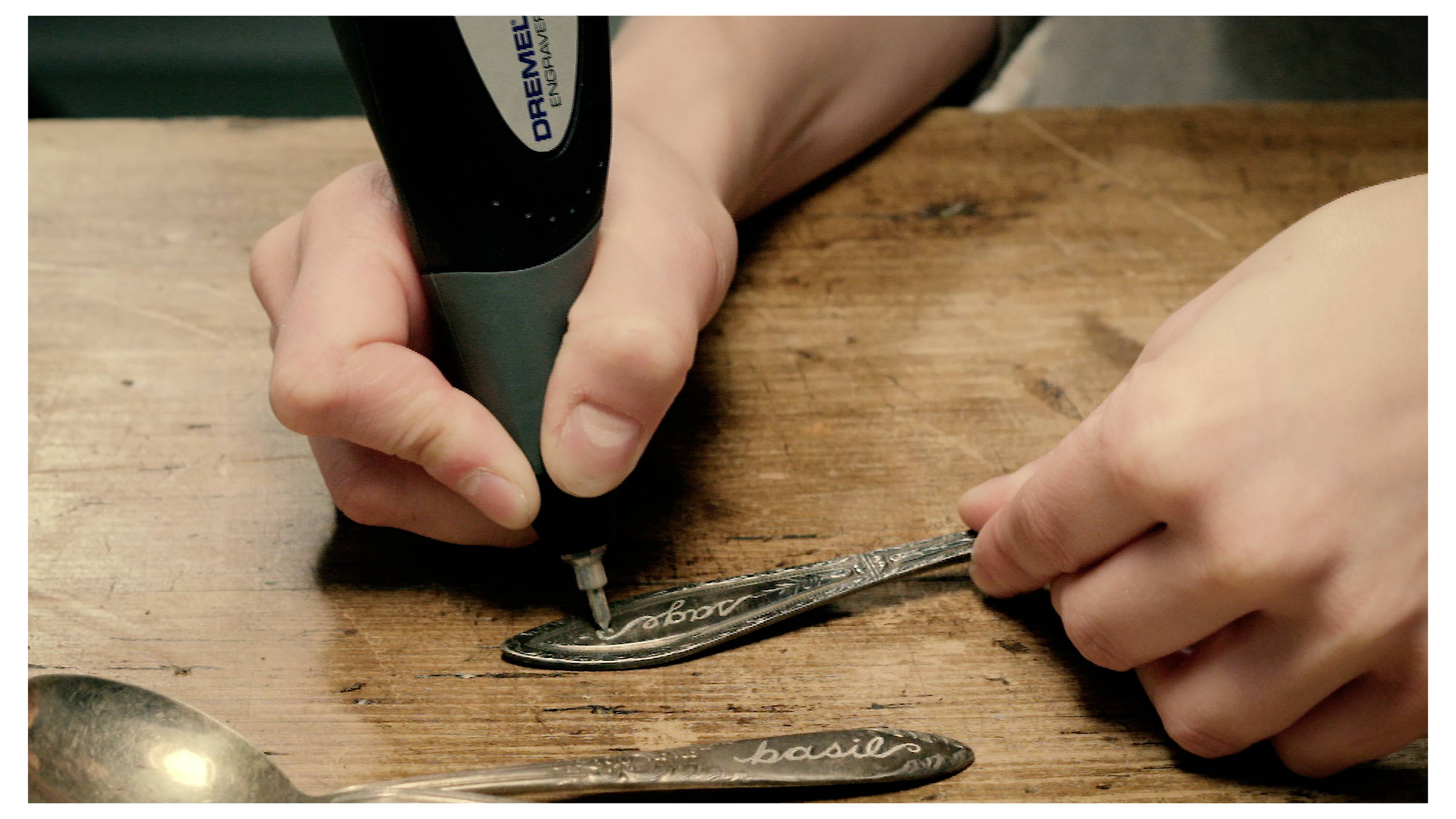 Osoba grawiruje słowo "rozmaryn" na łyżce za pomocą narzędzia do grawerowania, a obok na drewnianym stole znajduje się inna łyżka oznaczona napisem "bazylia".