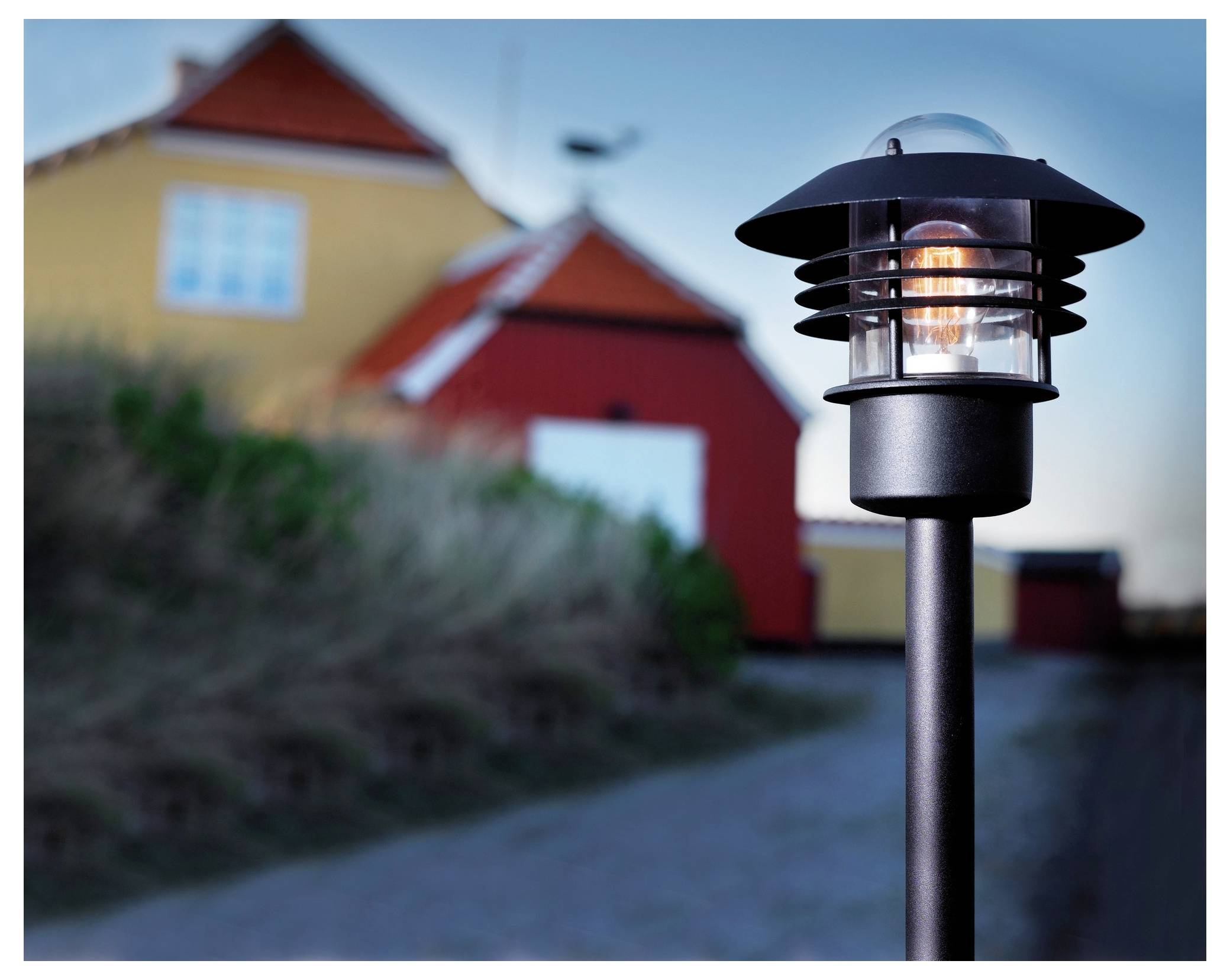 Oświetlony zewnętrzny słupek lampy znajduje się na pierwszym planie z rozmytymi żółtymi i czerwonymi domami w tle, tworząc przytulną atmosferę zmierzchu.