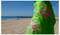 Osoba ubrana w jasno zieloną koszulkę z różowymi wzorami morskich żółwi stoi na piaszczystej plaży z oceanem i drewnianym molem w tle.