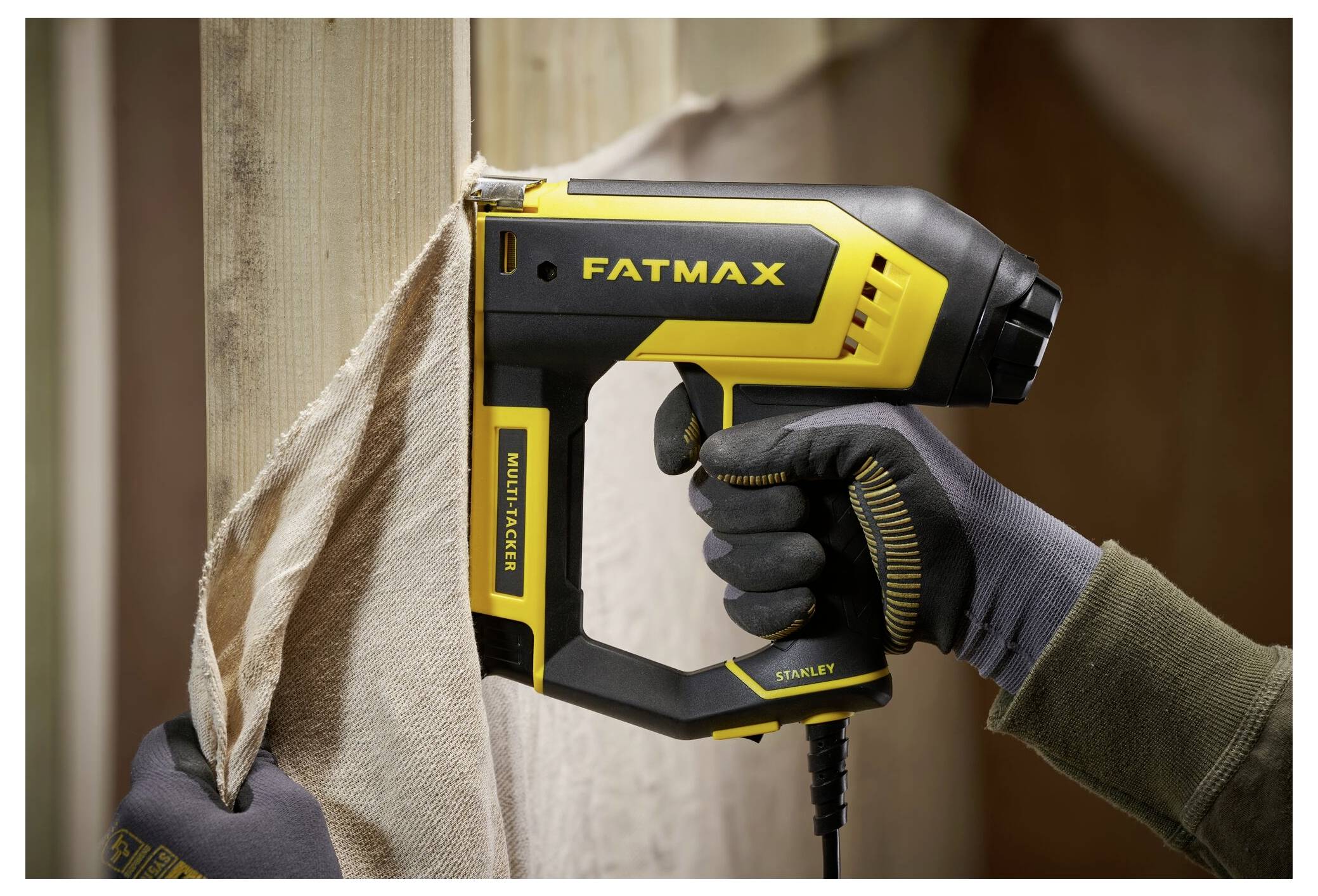 Osoba w rękawiczkach używa żółto-czarnego zszywacza do tkanin oznaczonego 'FATMAX' w celu przymocowania materiału do drewnianej ramy.