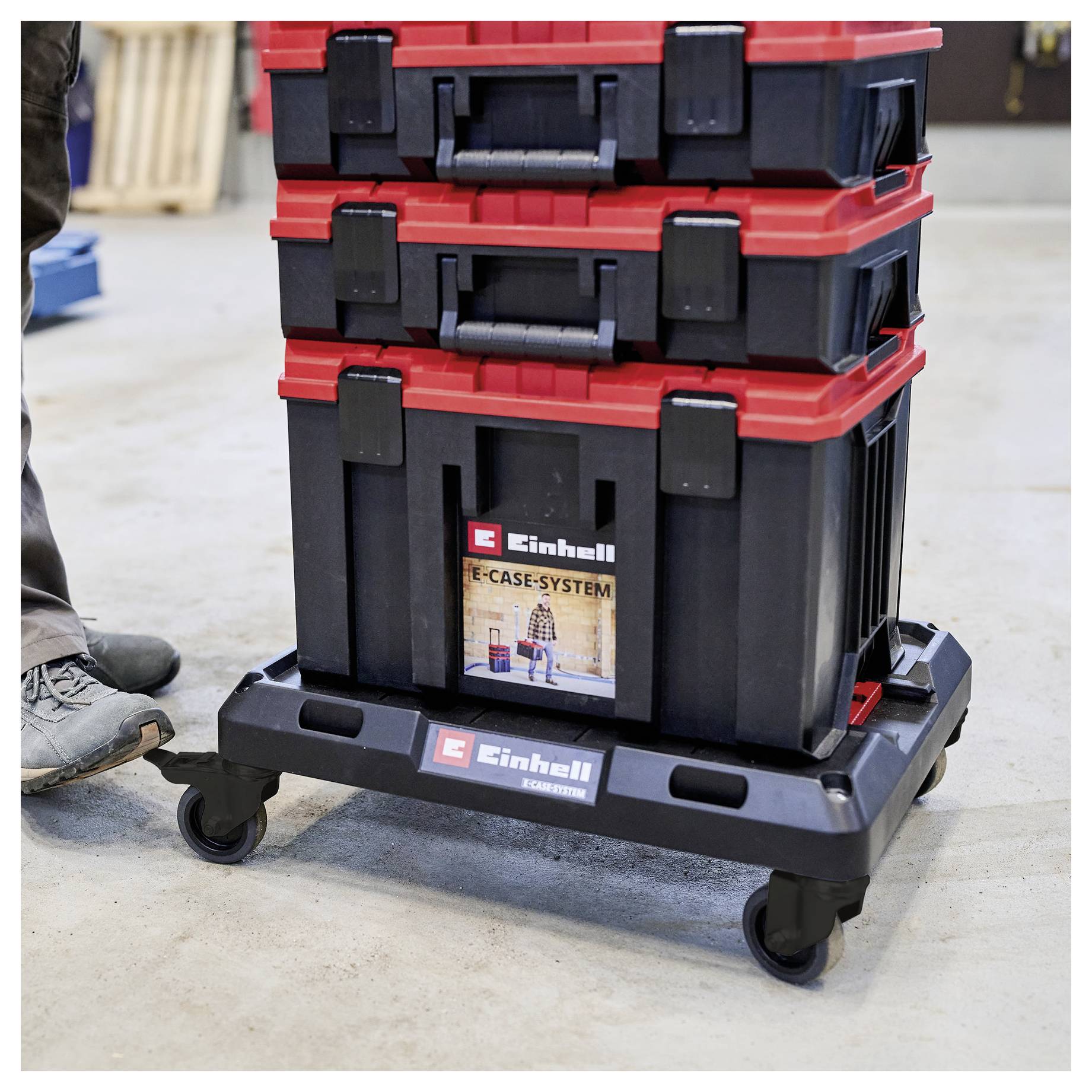 Ułożone w stos skrzynki narzędziowe Einhell na wózku z kółkami w warsztacie, z częściowo widocznym butem osoby po lewej stronie.