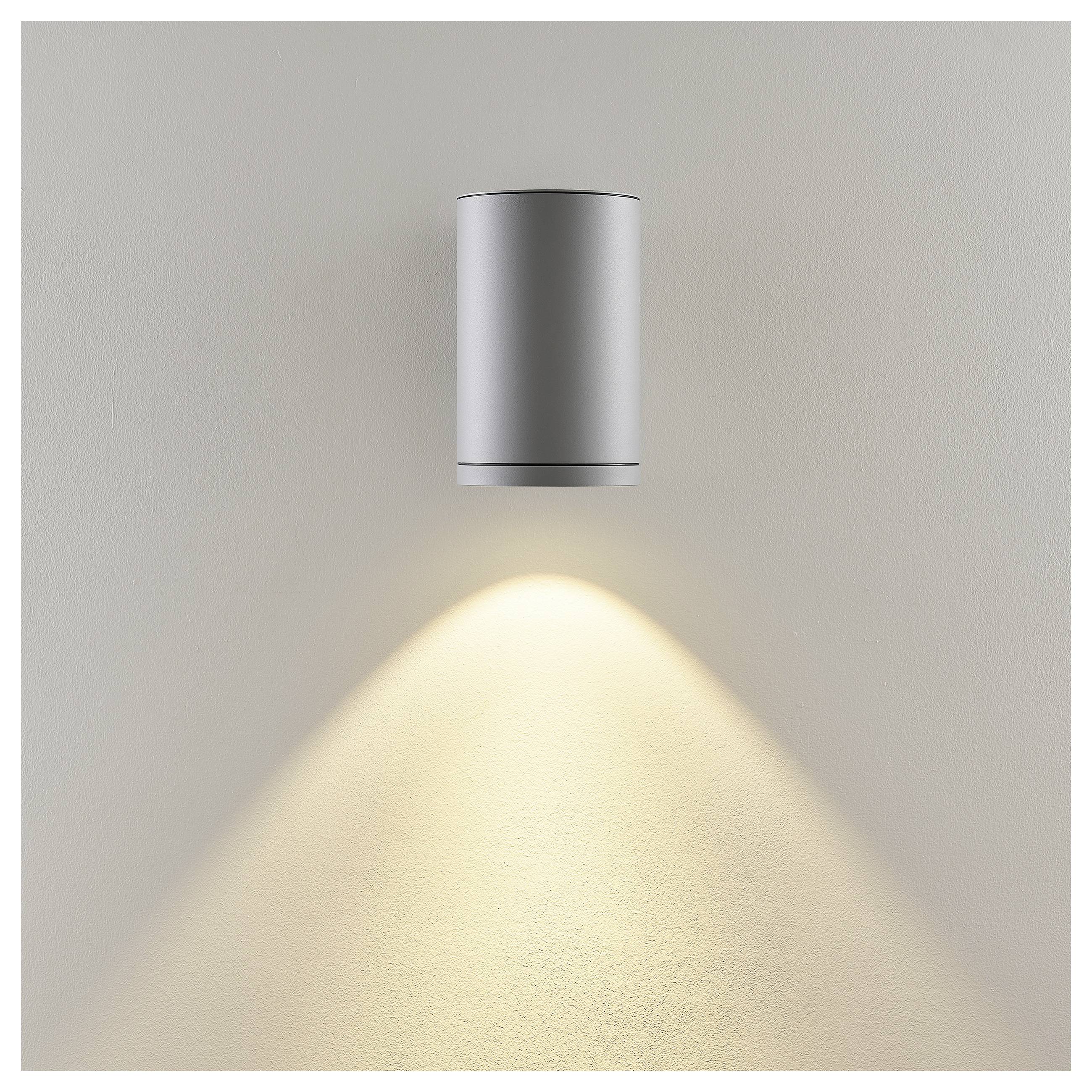 Ścienna srebrna cylindryczna lampa oświetleniowa rzucająca ciepłe światło w dół, tworząc stożek światła na zwykłej beżowej ścianie.