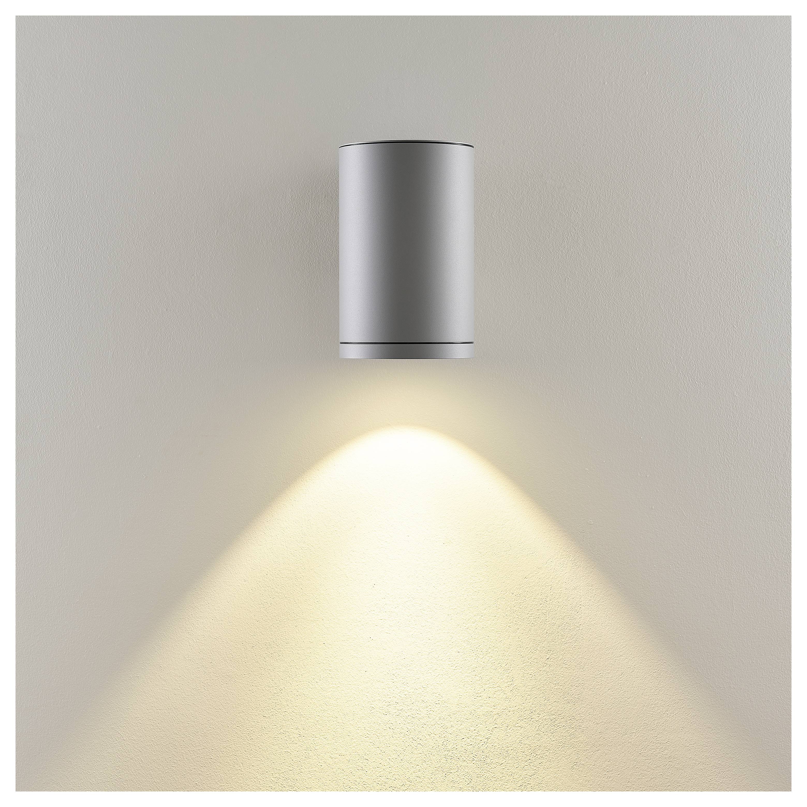 Ścienna srebrna cylindryczna lampa rzucająca ciepły stożek światła na beżową ścianę.
