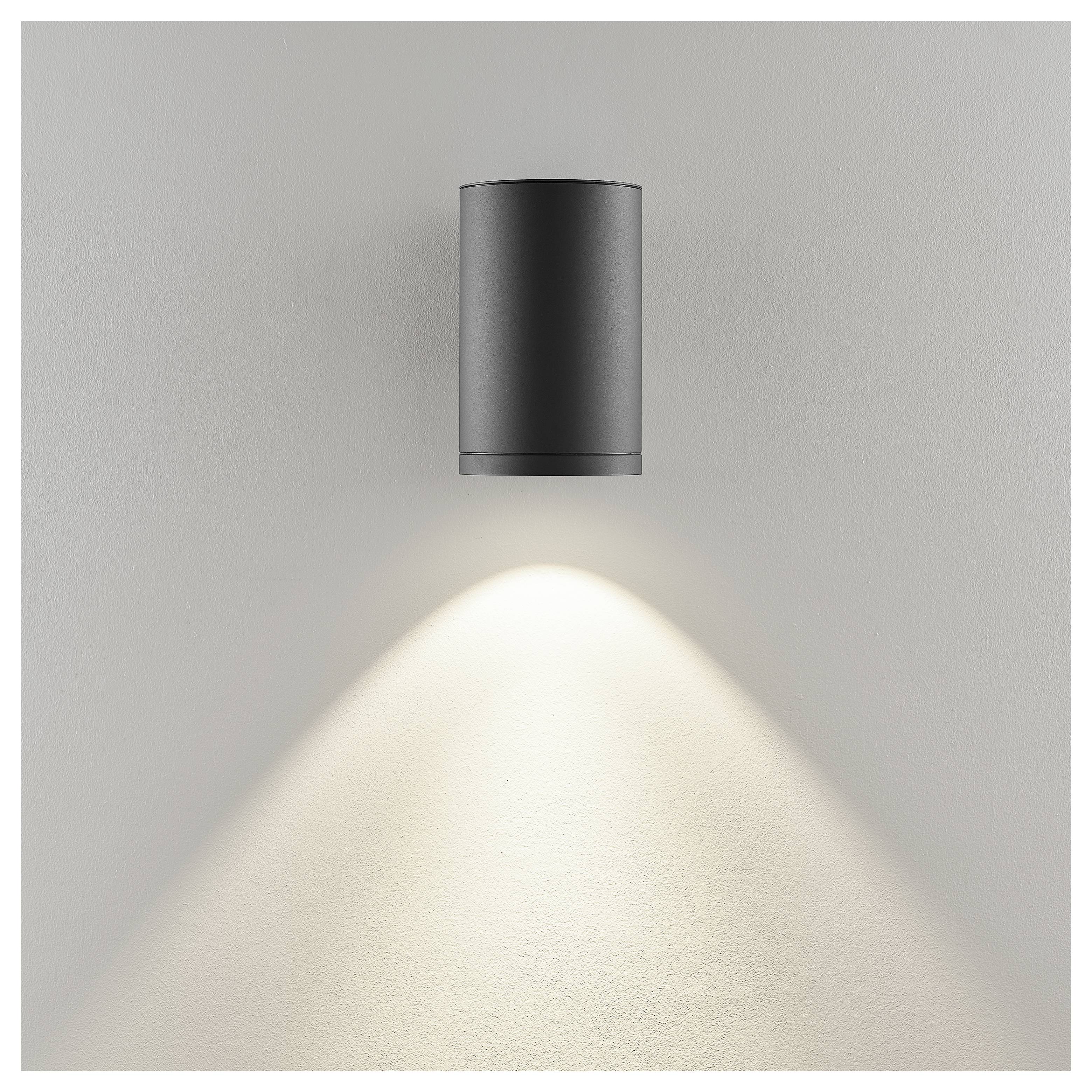 Czarna cylindryczna lampa ścienna na szarej ścianie, rzucająca ciepły stożek światła w dół.