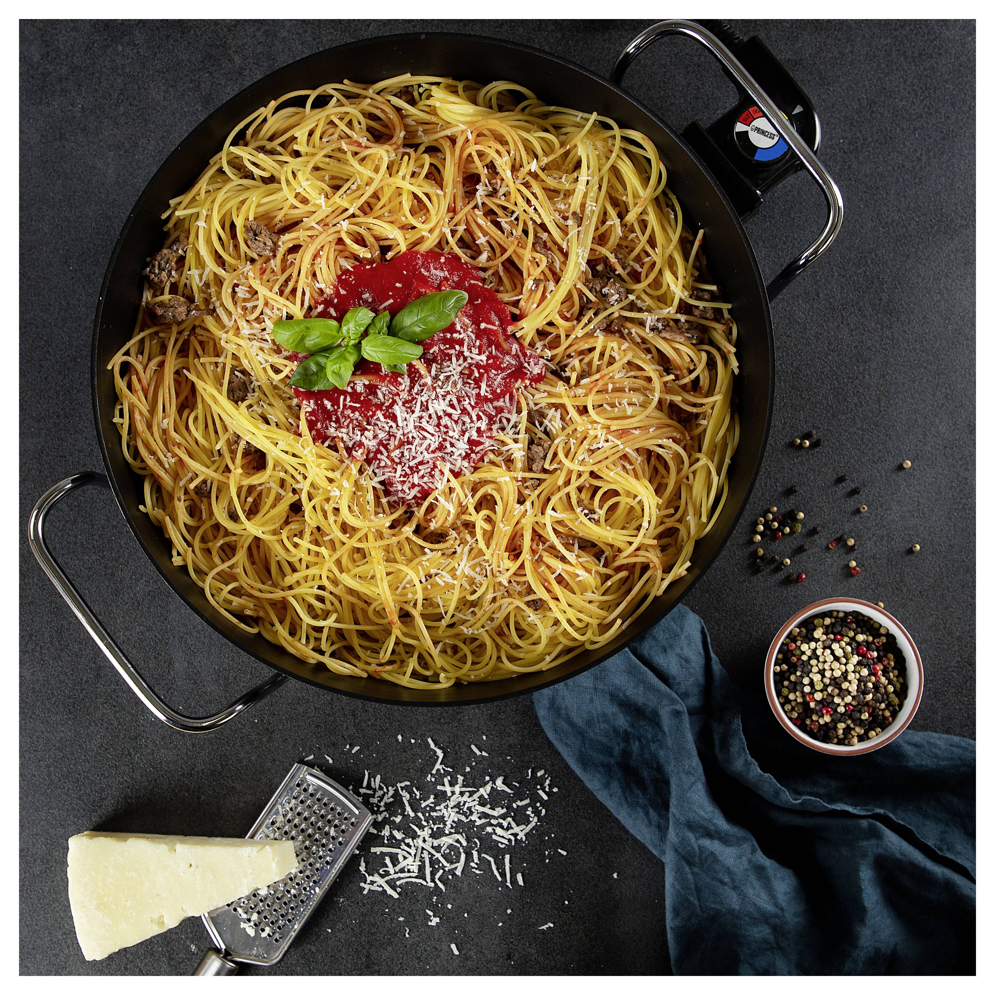 Zdjęcie z góry makaronu spaghetti w czarnej patelni z sosem pomidorowym, bazylią i startym serem, z kawałkiem sera, tarką, ziarnami pieprzu i tkaniną w pobliżu.