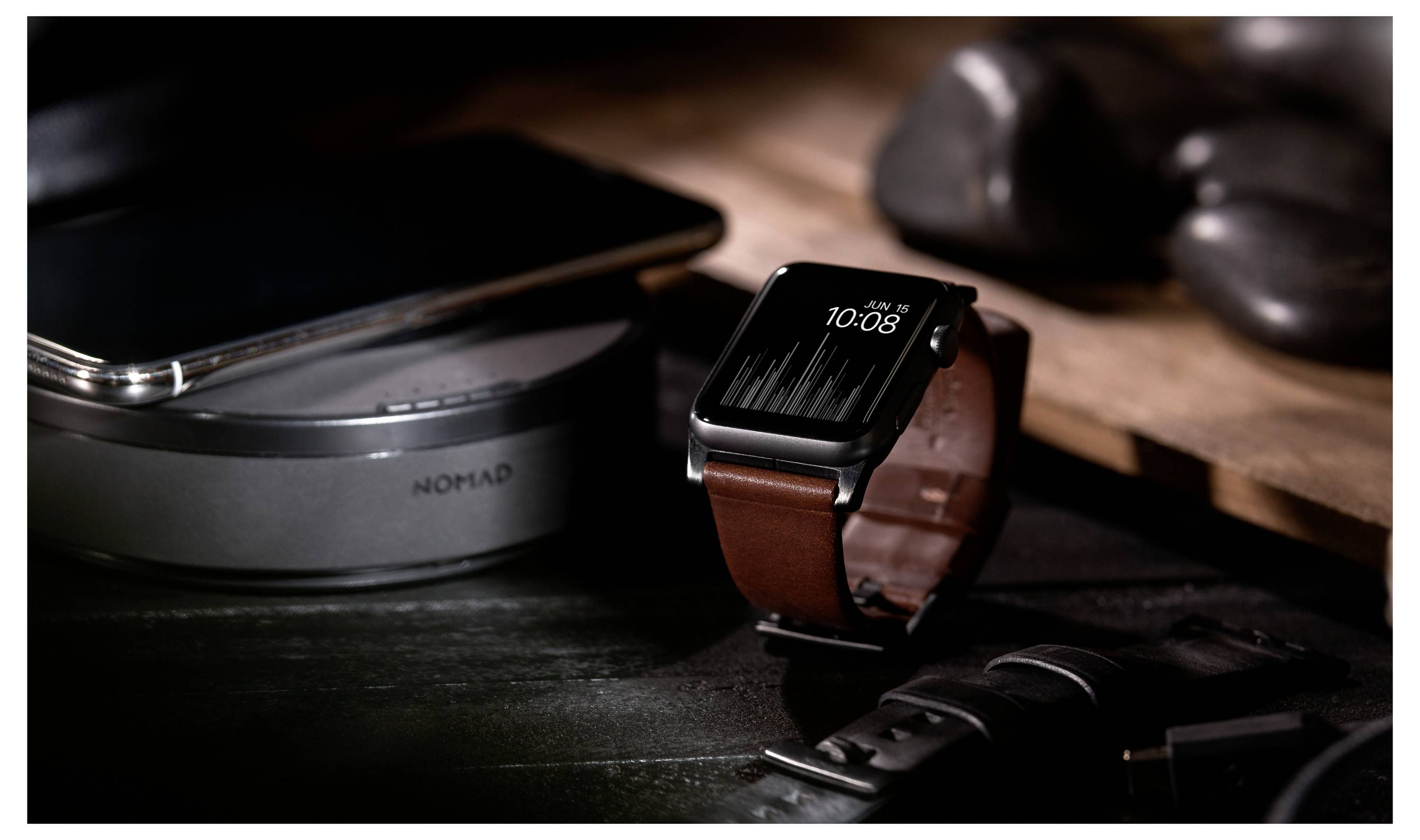 Smartwatch z brązowym skórzanym paskiem na biurku, ładujący się obok telefonu na bezprzewodowej stacji Nomad, z kablami i akcesoriami w pobliżu.