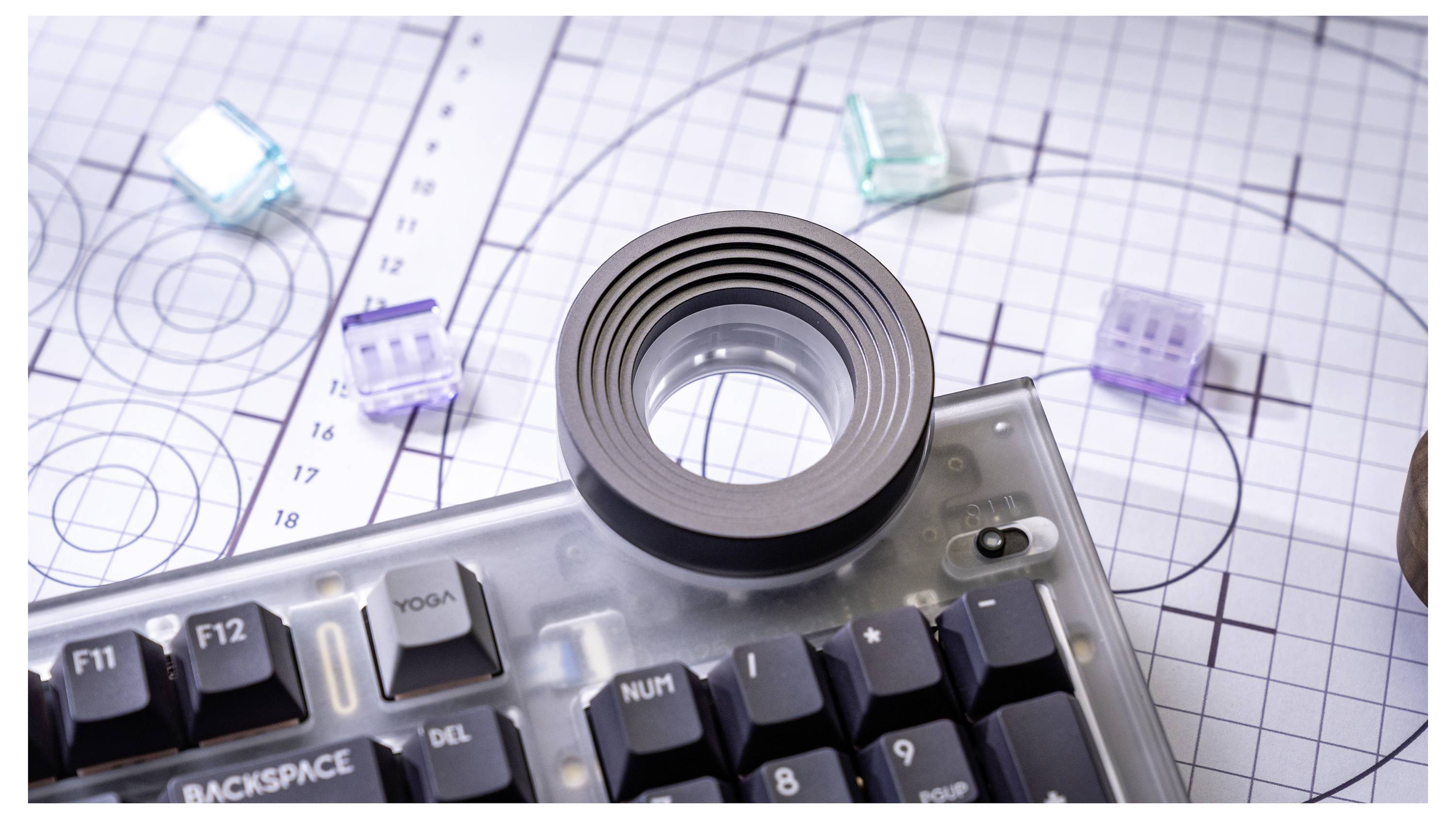 Zbliżenie mechanicznej klawiatury z lupą umieszczoną na wierzchu, położone na papierze milimetrowym z diagramami przypominającymi obwody oraz małymi półprzezroczystymi sześcianami.