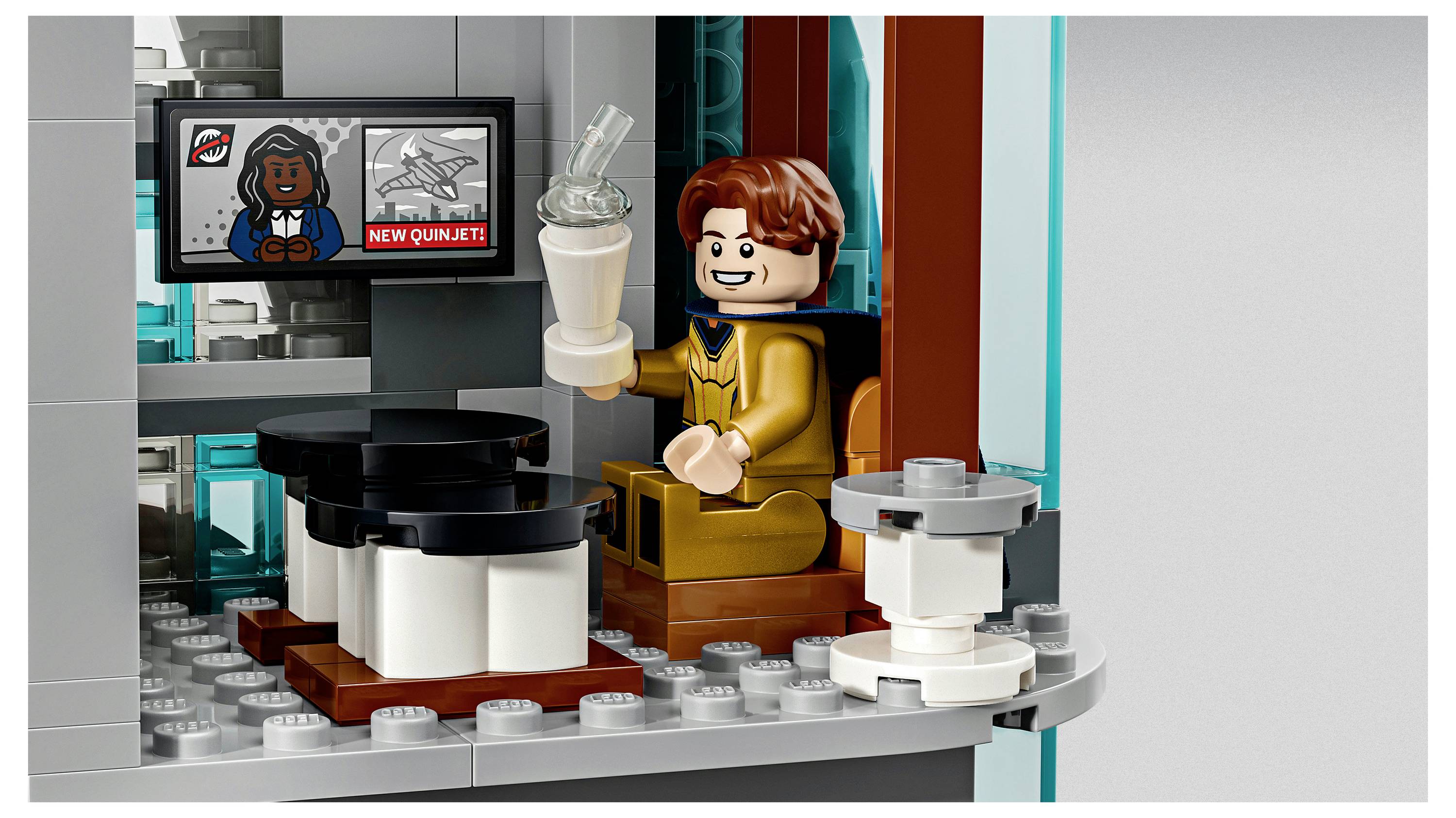 Minifigurka w stylu LEGO w złotym garniturze siedzi przy biurku, trzymając koktajl mleczny i patrzy na monitor, na którym wyświetla się postać z kreskówki i tekst „NOWY QUINJET".