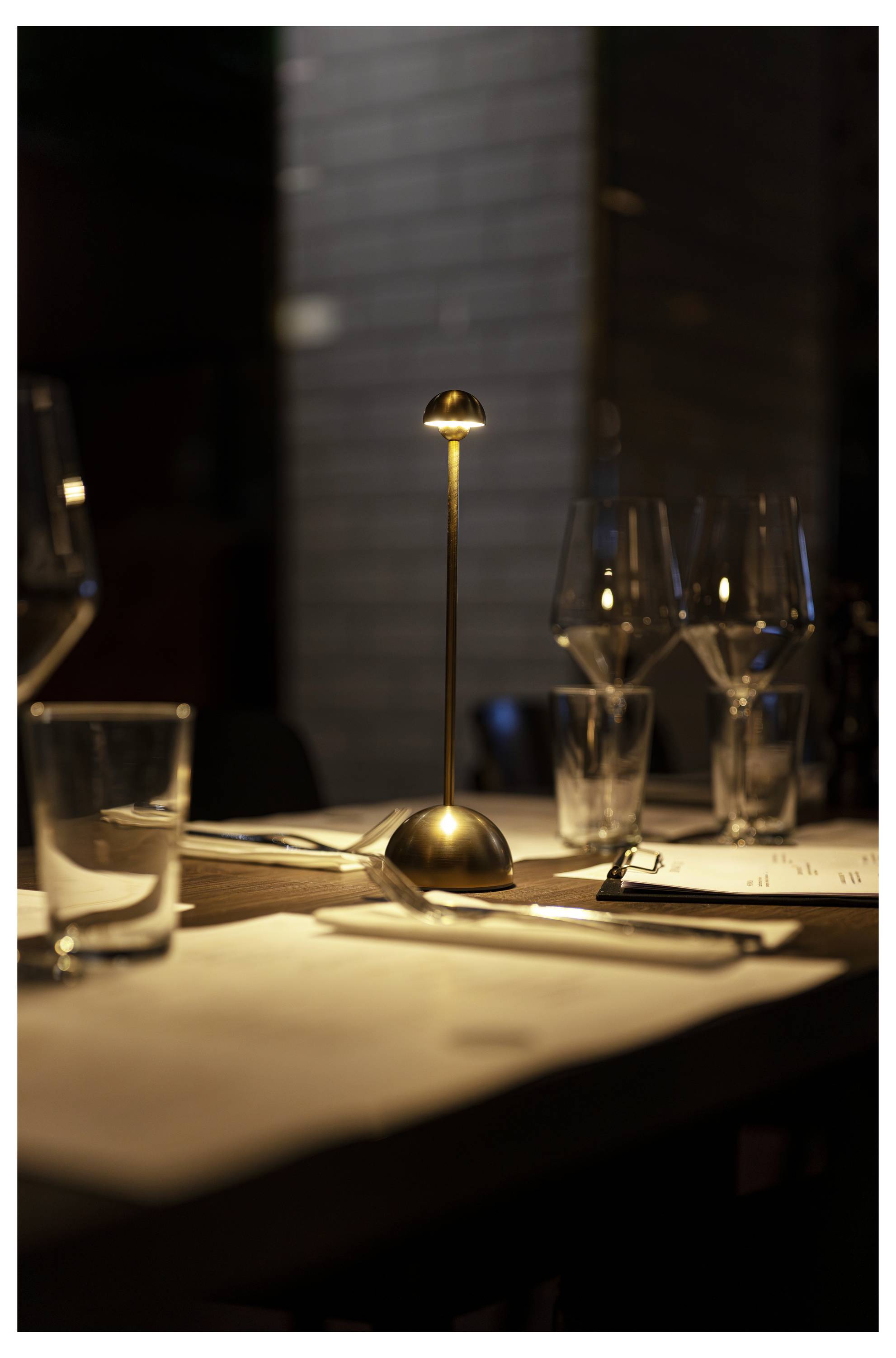 Stół w restauracji oświetlonej świecami, zastawiony kieliszkami, sztućcami, menu i małą lampką, tworzący eleganckie ustawienie do kolacji.