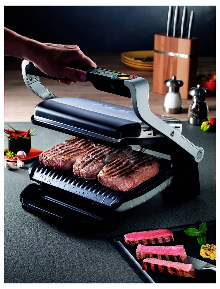 Ręka naciskająca grill blat kuchenny zamykając go nad syczącymi stekami w kuchni, z pokrojonymi grillowanymi mięsem podawanym na czarnym talerzu.