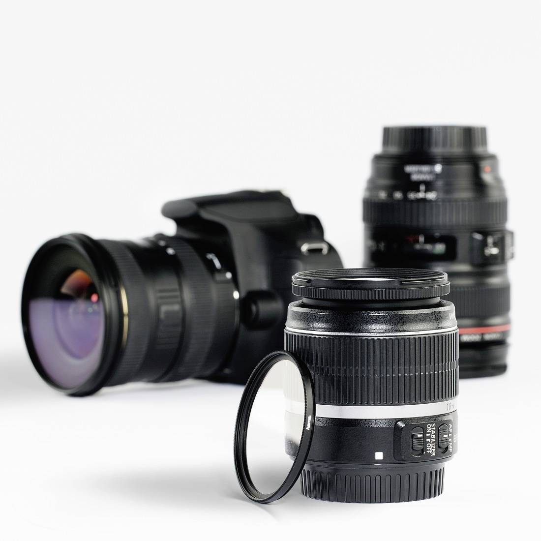 Trzy obiektywy do aparatu fotograficznego i aparat DSLR na białym tle, z filtrem do obiektywu stojącym z przodu.