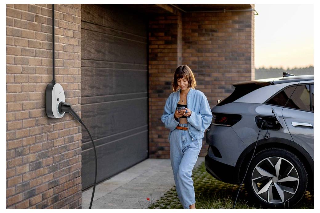 Kobieta używająca smartfona stojąc obok samochodu elektrycznego podłączonego do ładowarki ściennej poza murowanym garażem.