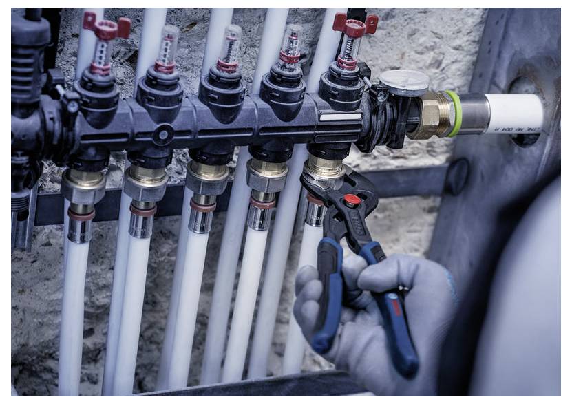 Pracownik w rękawiczkach regulujący zawór na kolektorze z kilkoma białymi rurami plastikowymi w przemysłowym systemie hydraulicznym.