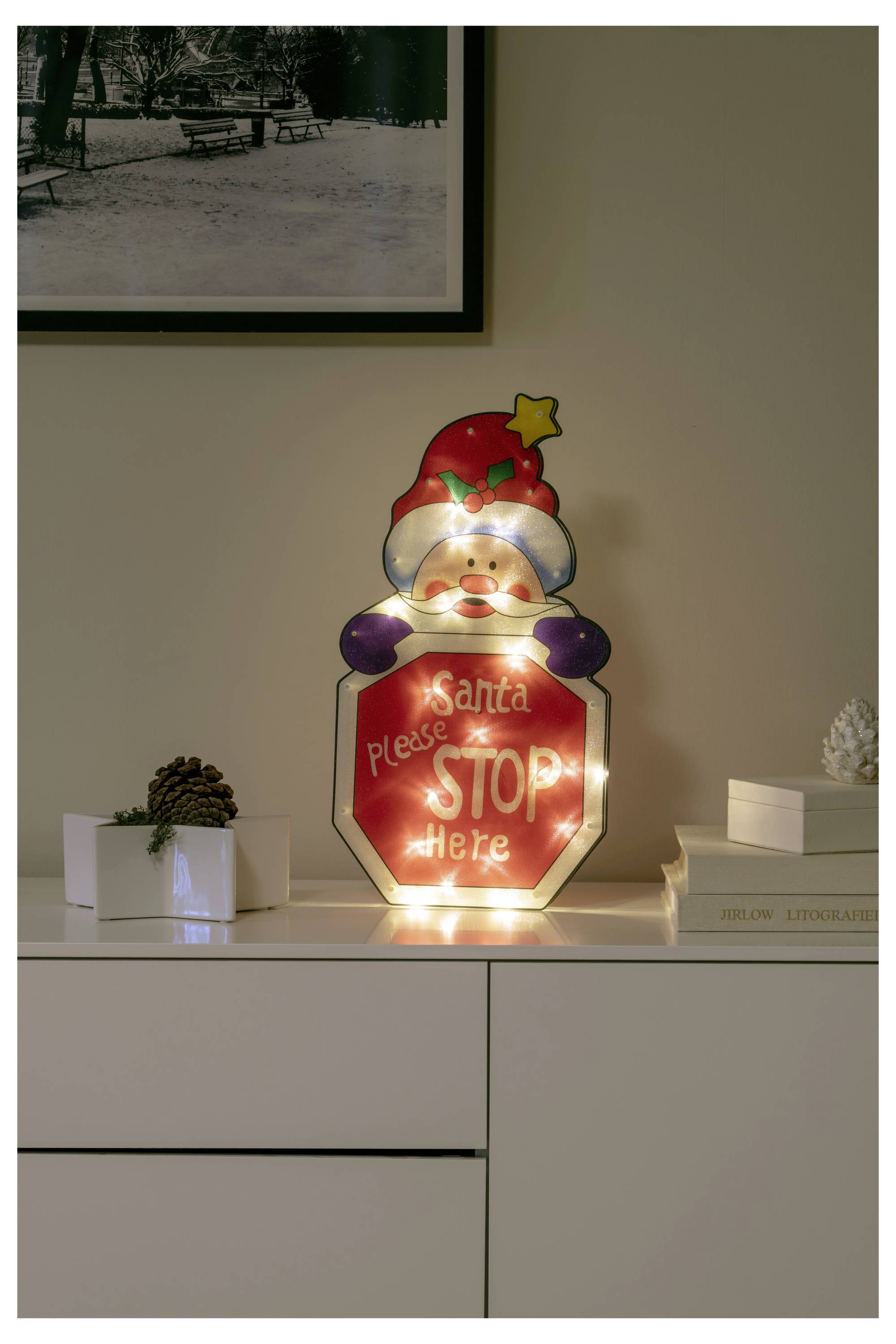 Podświetlony znak w kształcie Świętego Mikołaja na komodzie z napisem „Santa please STOP here", obok małej rośliny ze szyszkami i książek.