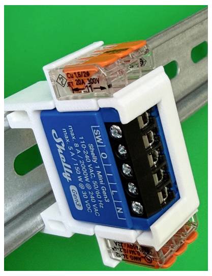 Zbliżenie niebieskiego modułu przekaźnika elektrycznego montowanego na szynie DIN z przezroczystymi złączami zaciśniętymi do metalowej szyny na zielonym tle.