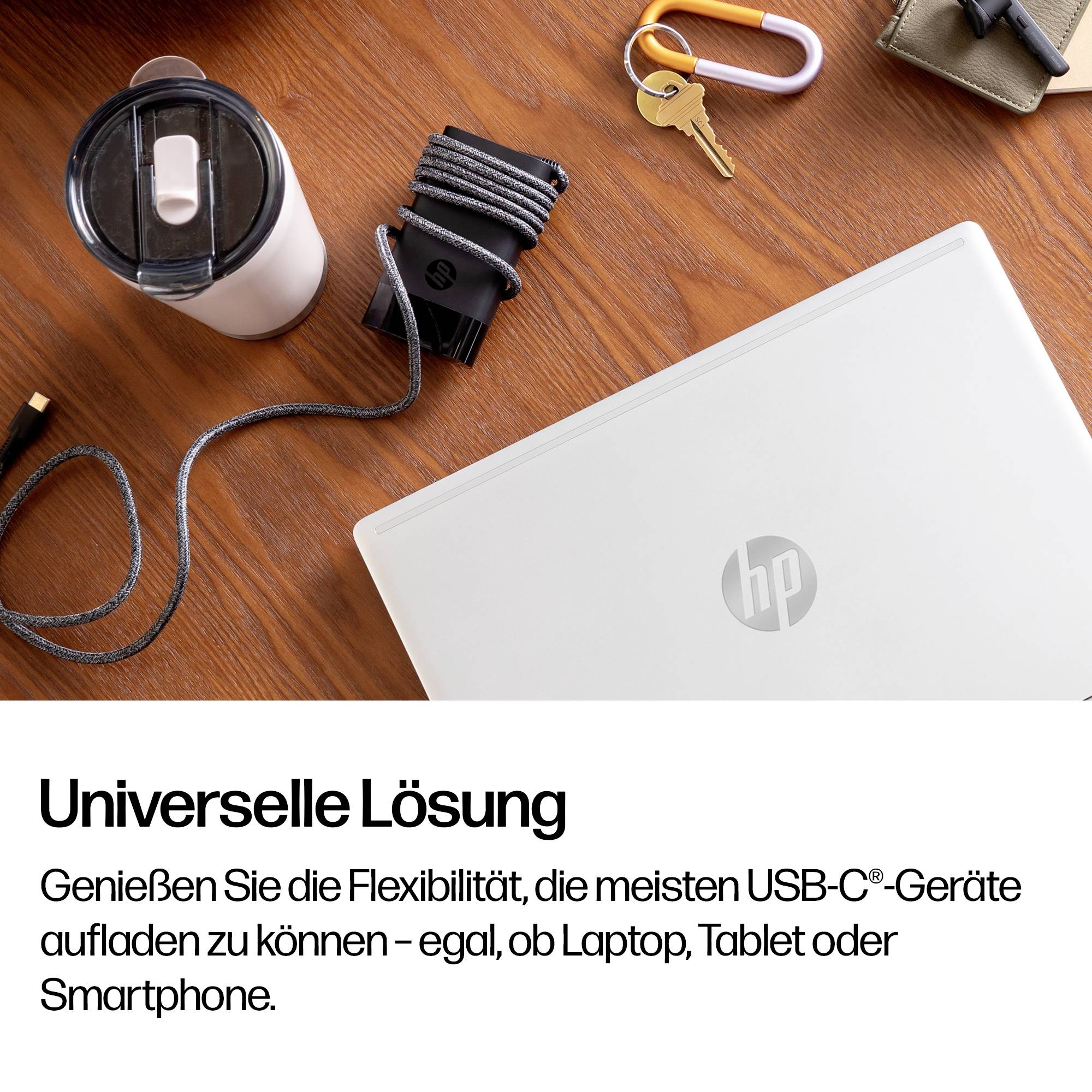 Laptop HP z ładowarką USB-C, kabel, butelka na wodę i klucze na biurku; tekst niemiecki mówi „Uniwersalne Rozwiązanie" i promuje ładowanie większości urządzeń USB-C.