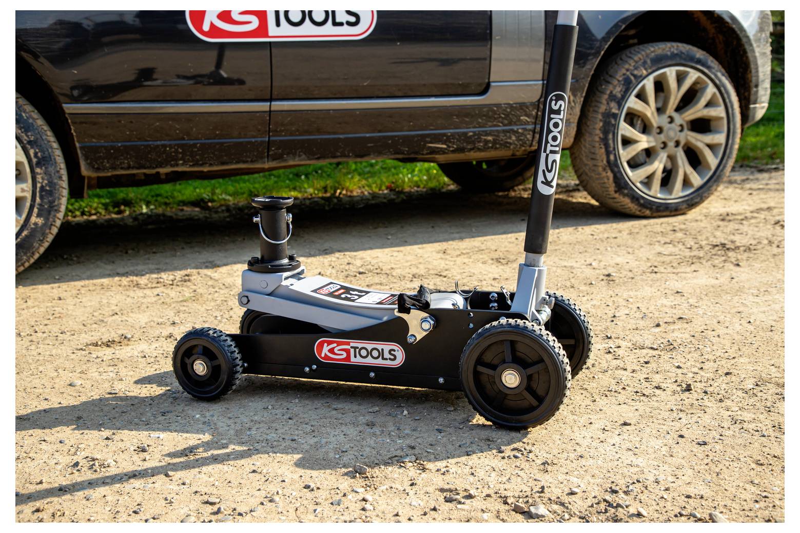 Niskobudżetowy czarny hydrauliczny podnośnik podłogowy KS Tools na żwirowej powierzchni obok SUV-a z widocznym podniesiony uchwyt do podnoszenia.