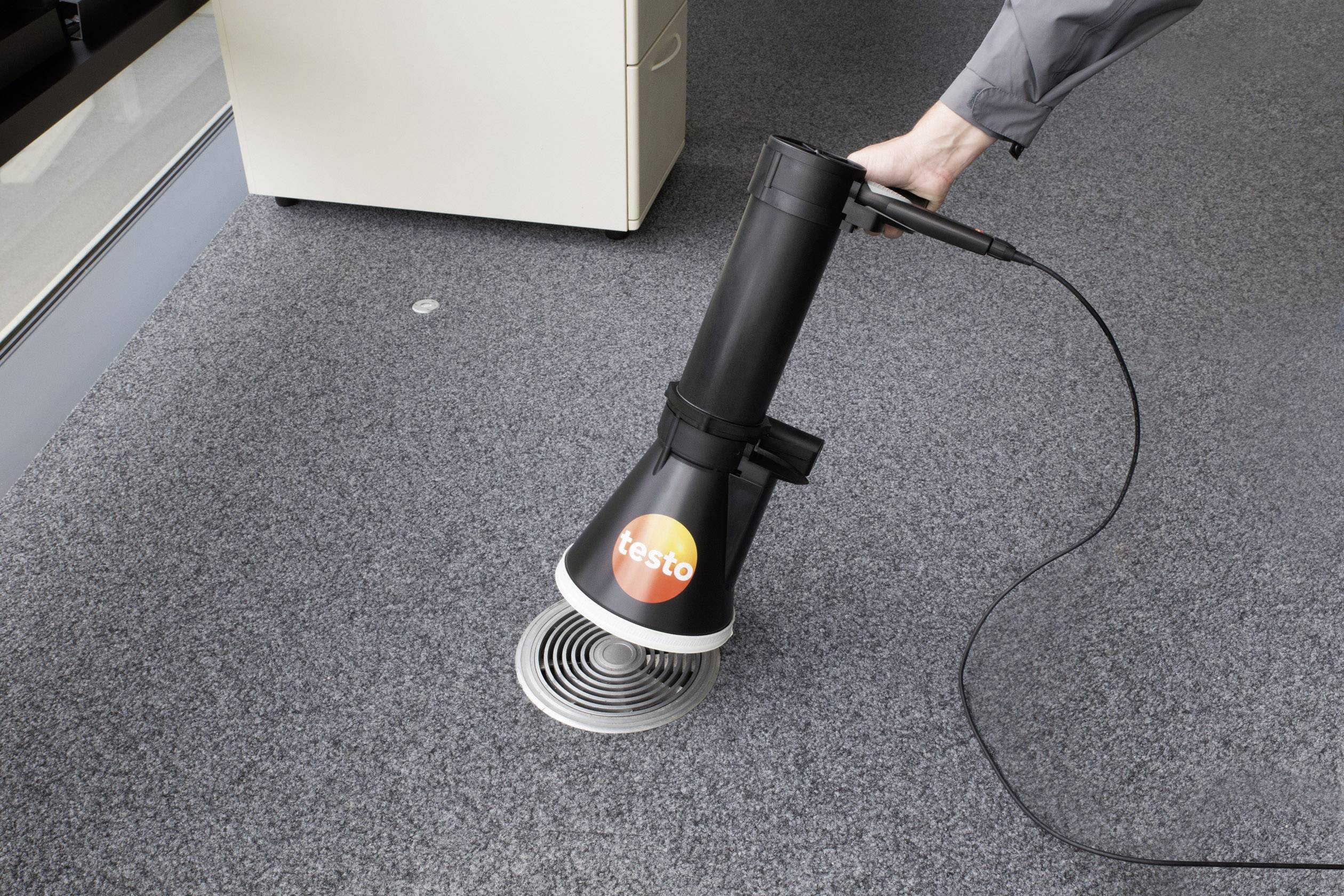 Osoba trzyma czarny przyrząd pomiarowy nad okrągłym otworem wentylacyjnym na szarym dywanie.