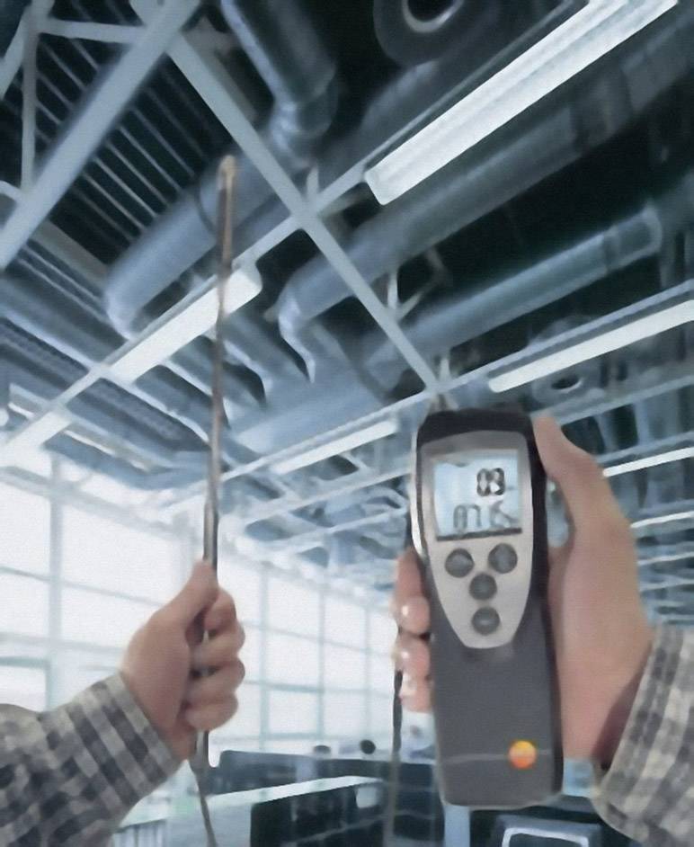 Osoba trzyma w ręku miernik przepływu powietrza, mierząc strumień powietrza w przemysłowym systemie wentylacyjnym z dużymi przewodami.