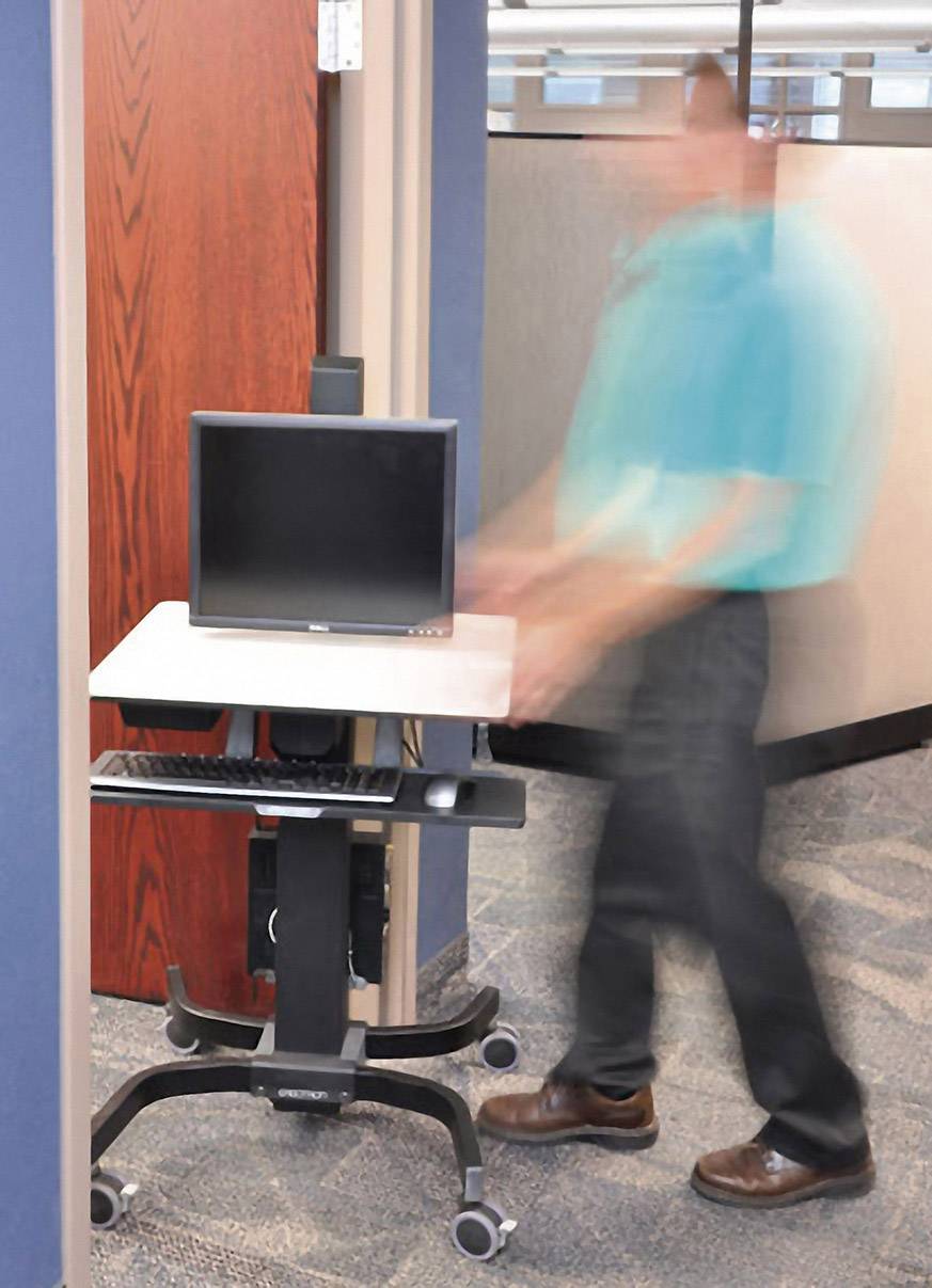 Mężczyzna przesuwa mobilny stolik komputerowy przez drzwi biurowe. Na stoliku znajduje się monitor i klawiatura.