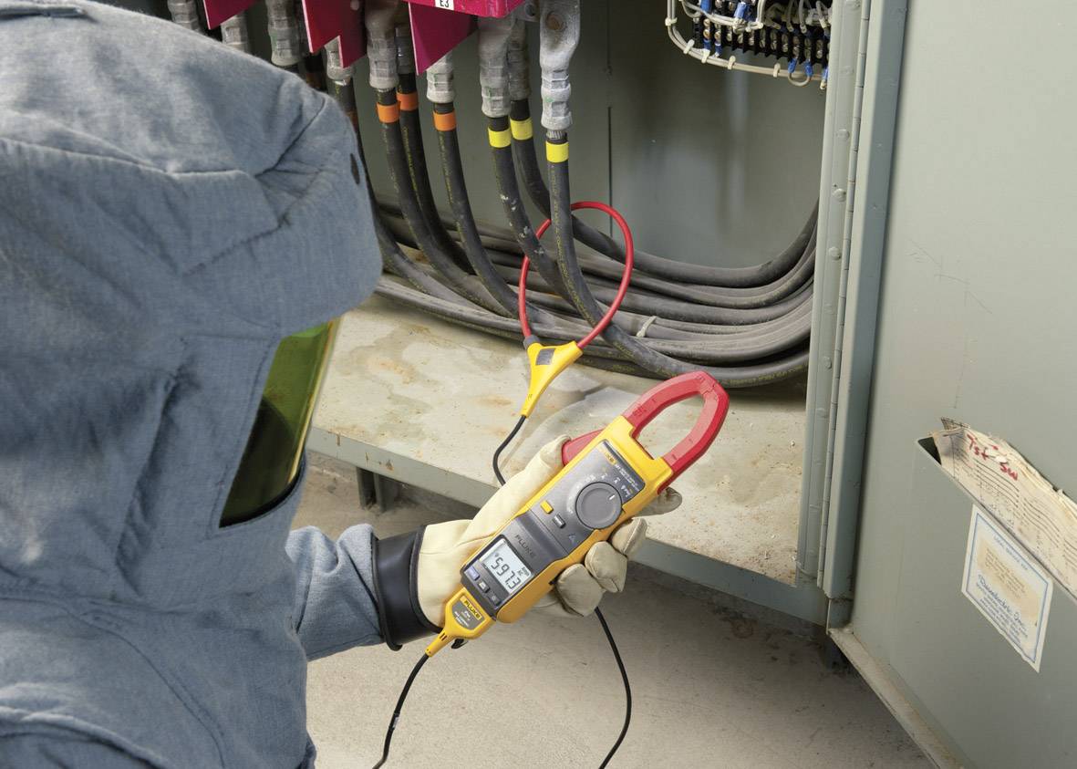 Osoba w odzieży ochronnej mierzy napięcie elektrycznej rozdzielni za pomocą cyfrowego multimetru. Przestrzegane są środki bezpieczeństwa.