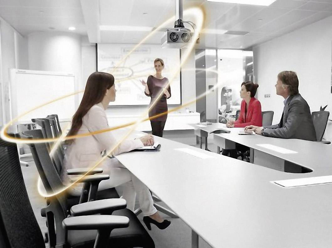 Osoby w sali konferencyjnej, kobieta stoi i przemawia, inni słuchają. Smugę świetlną symbolizuje komunikacja lub innowacja.