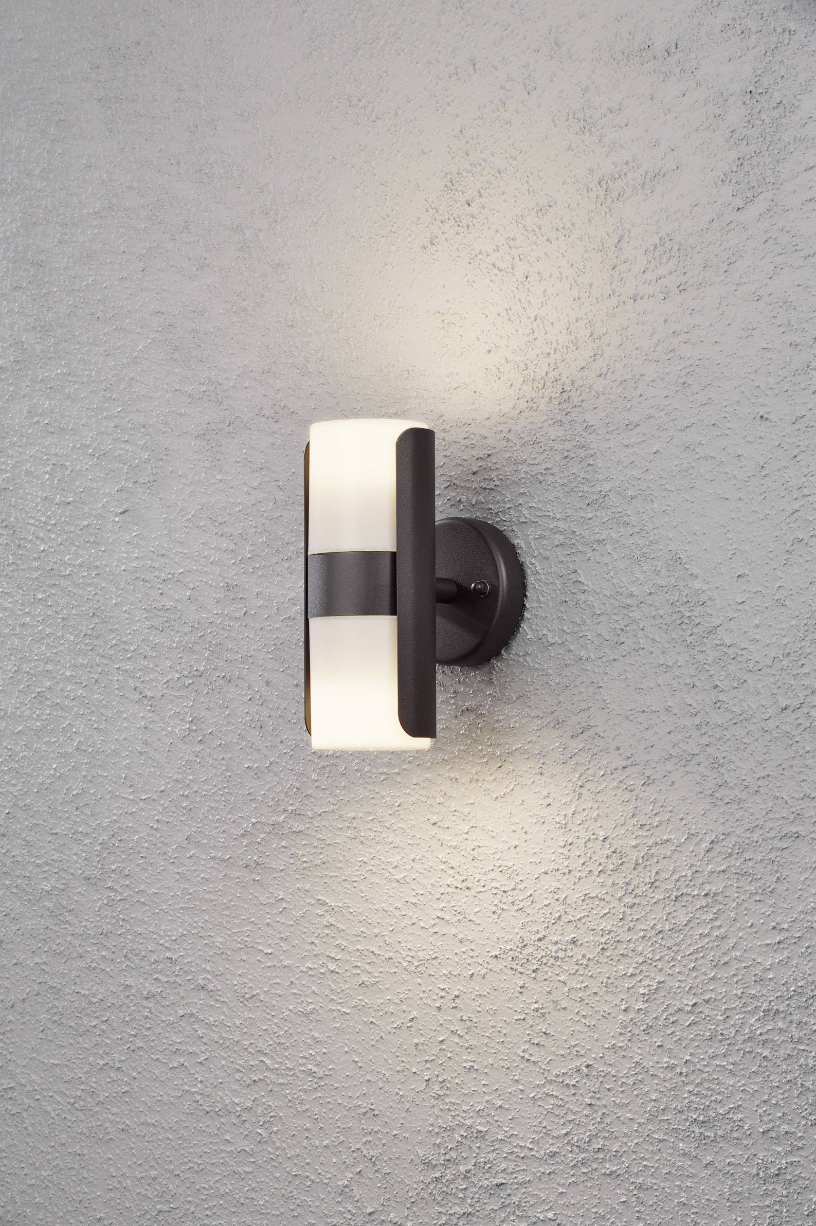 Kinkiet o nowoczesnym designie, wykonany z czarnego metalu i mlecznego szkła. Emituje ciepłe światło i jest zamontowany na szarej ścianie.