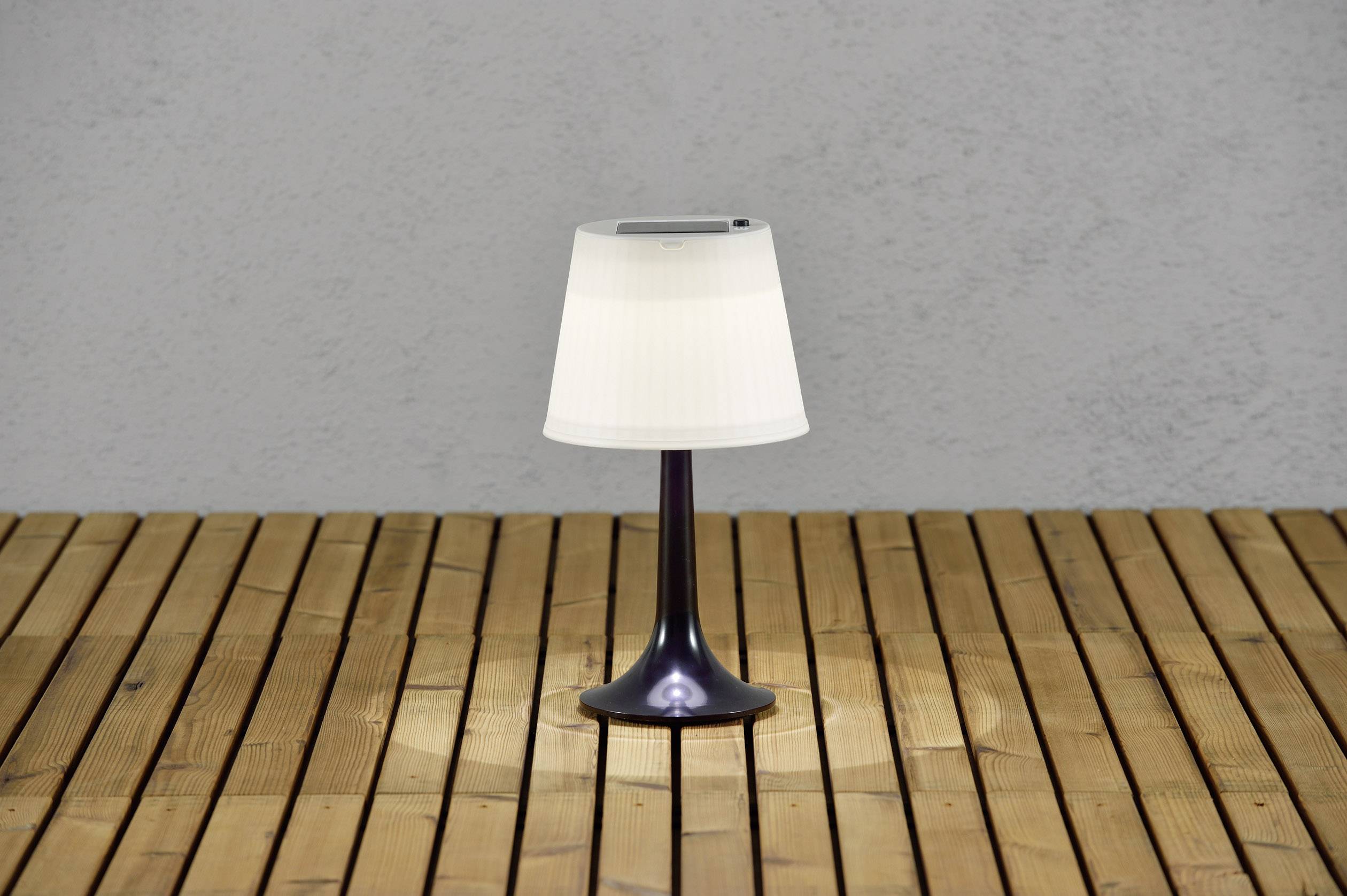 Pojedyncza lampa stołowa z białym abażurem stoi na drewnianej podłodze przed szarą ścianą.