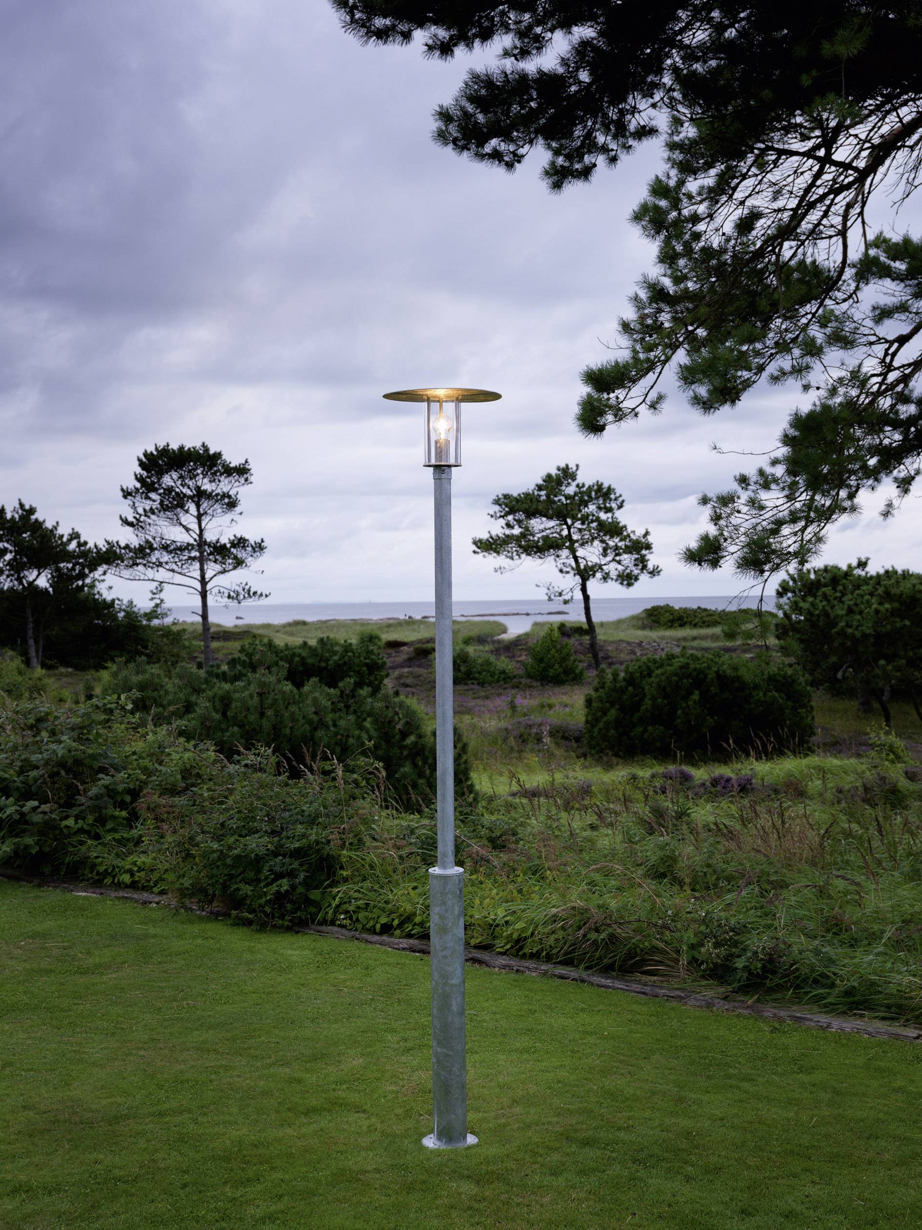 Latarnia uliczna stoi na łące przed porośniętym krajobrazem z drzewami i pochmurnym niebem w tle.