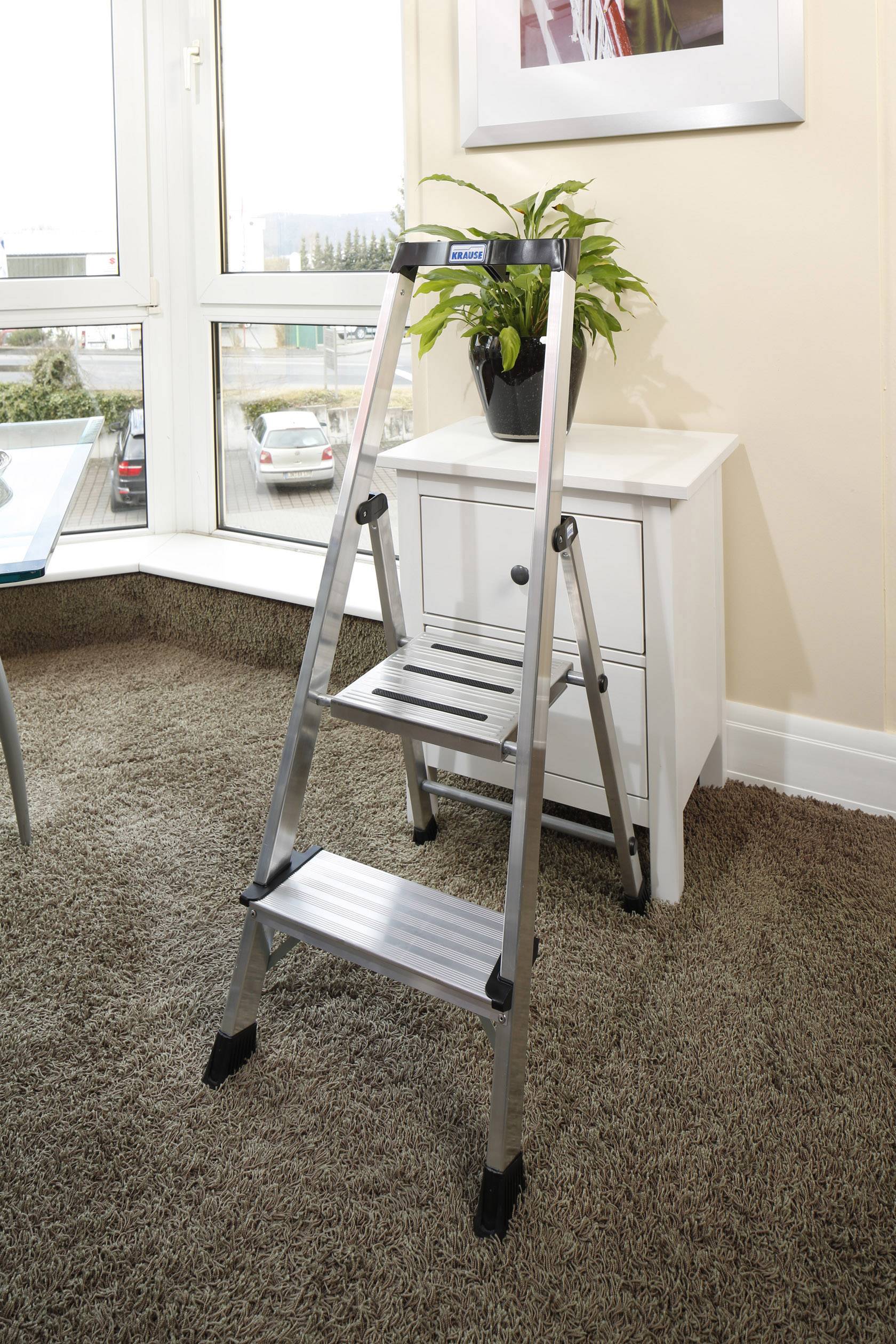 Metalowa drabina stoi na dywanie obok białej komody. Na komodzie znajduje się zielona roślina.