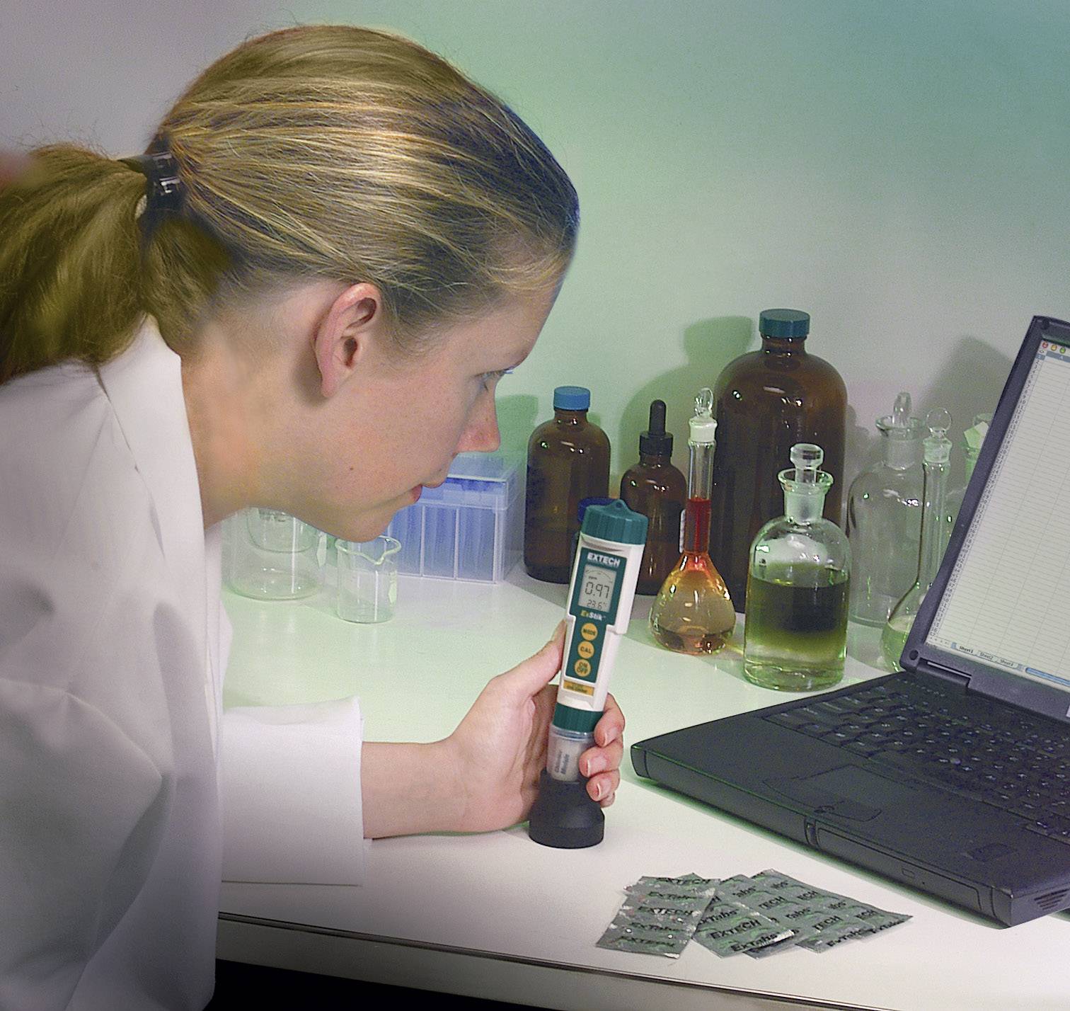 Kobieta w laboratorium mierzy pH płynu przenośnym urządzeniem, w otoczeniu butelek z chemikaliami i laptopa.