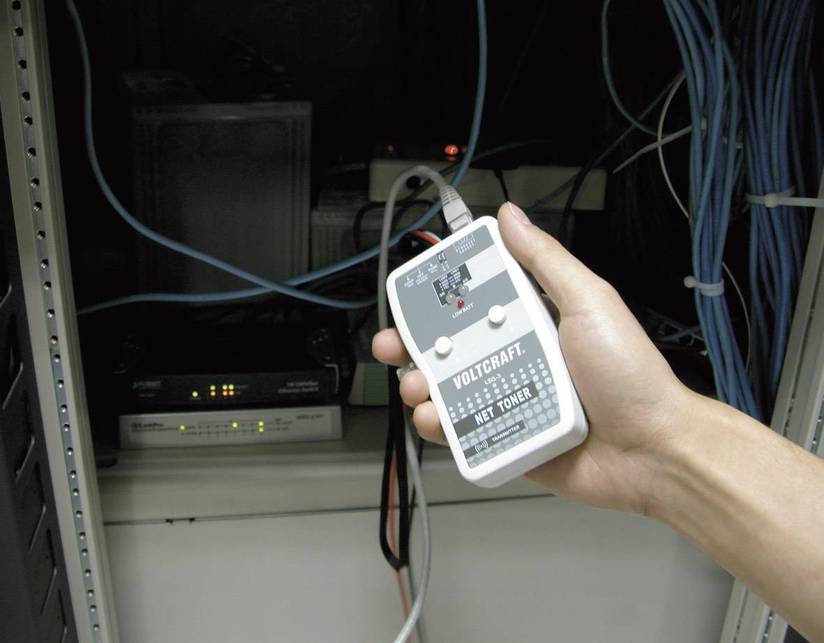 Ręka trzyma urządzenie do testowania kabli sieciowych w pomieszczeniu serwerowym. W tle widoczne są różne urządzenia elektroniczne i kable.