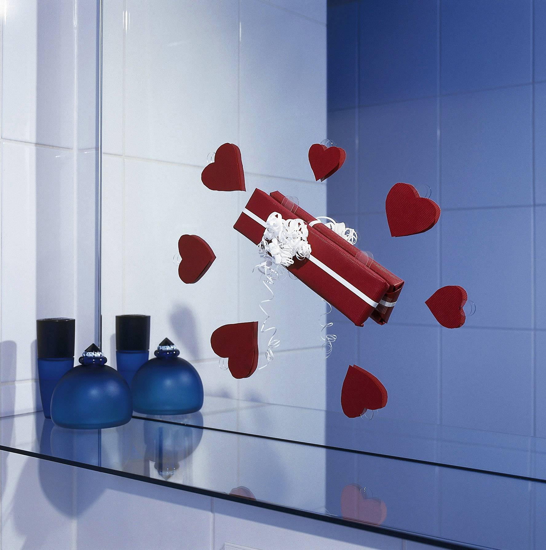 Czerwony prezent z białymi wstążkami unosi się otoczony czerwonymi sercami w niebieskim, kafelkowanym pomieszczeniu, obok stoją niebieskie butelki.