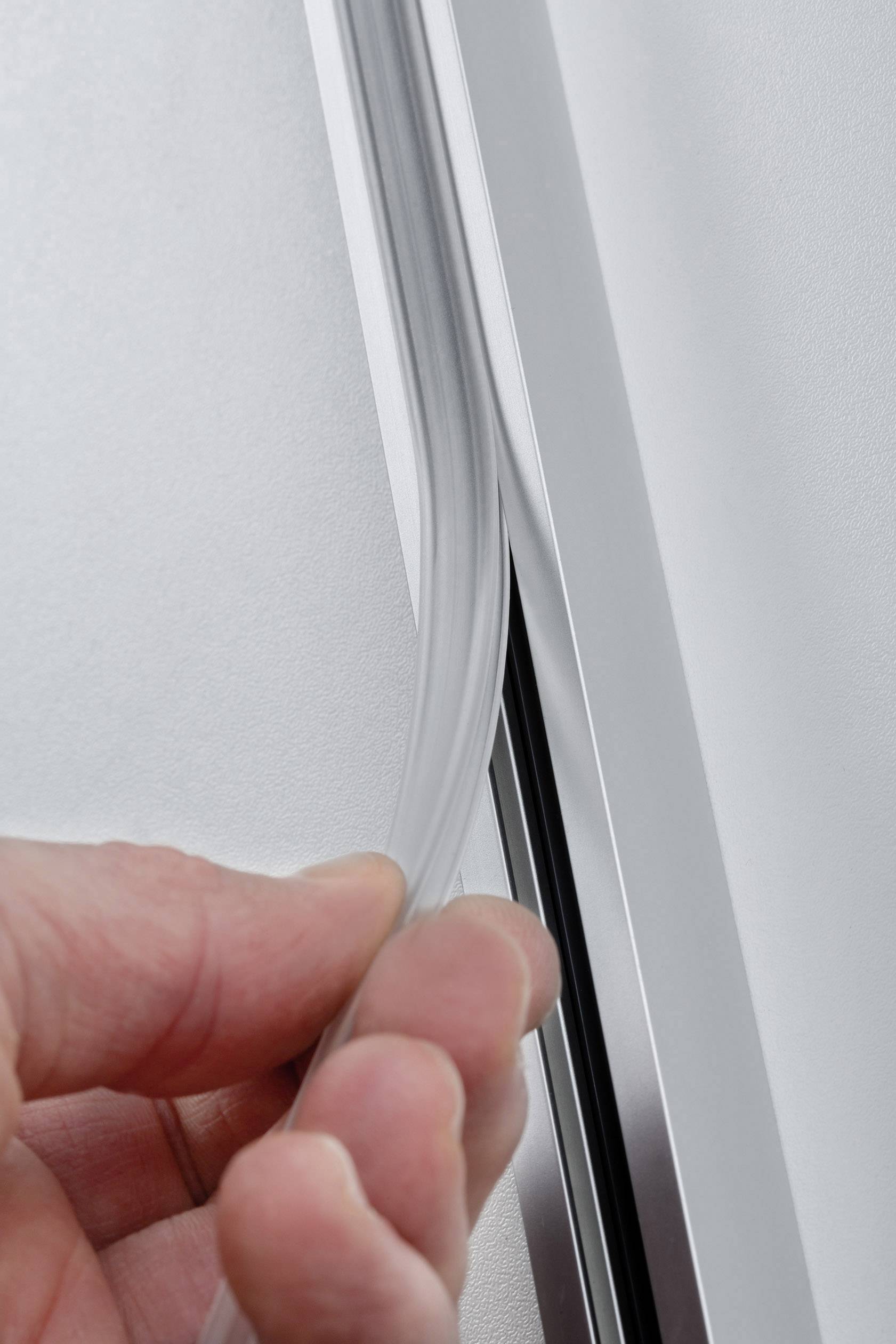 Ręka chwyta elastyczny, przezroczysty profil uszczelniający na srebrnej ramie. Służy do uszczelnienia lub ochrony.