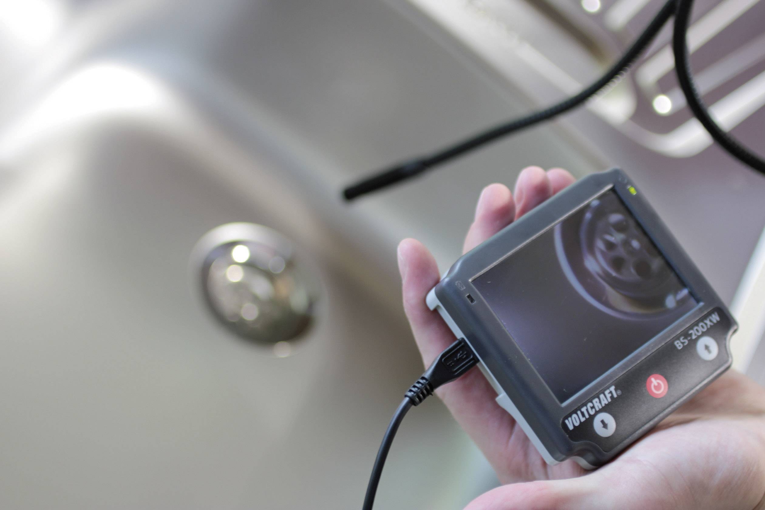 Dłoń trzyma urządzenie cyfrowe z ekranem, na którym widać powiększony obraz wnętrza rury. Na pierwszym planie widoczny jest zlew w tle.