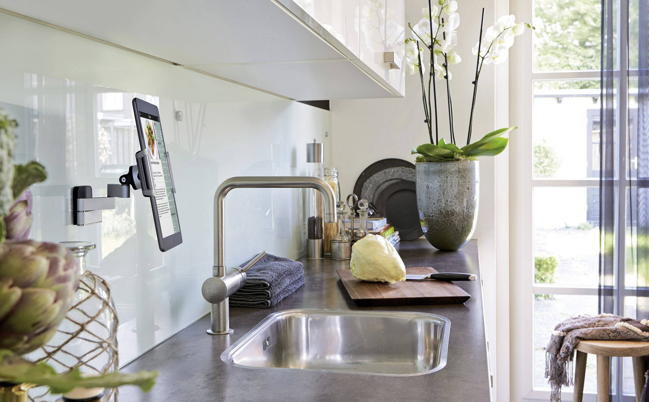 Nowoczesna kuchnia z zlewem, deską do krojenia i uchwytem na tablet. Storczyk stoi na parapecie. Jasna, przestronna atmosfera.