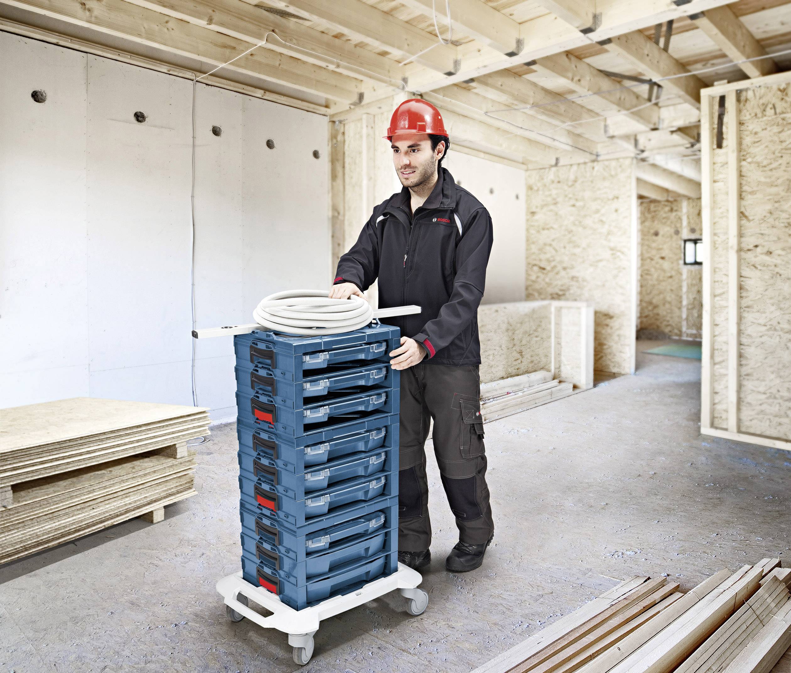 Robotnik w odzieży ochronnej i czerwonym kasku przesuwa wózek zułożonymi jedne na drugich skrzynkami w budynku w trakcie budowy drewnianej konstrukcji.
