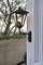 Lampa zewnętrzna z żarówką zamontowana jest na ścianie domu. W tle widoczny jest drewniany płot i nieostre drzewa.