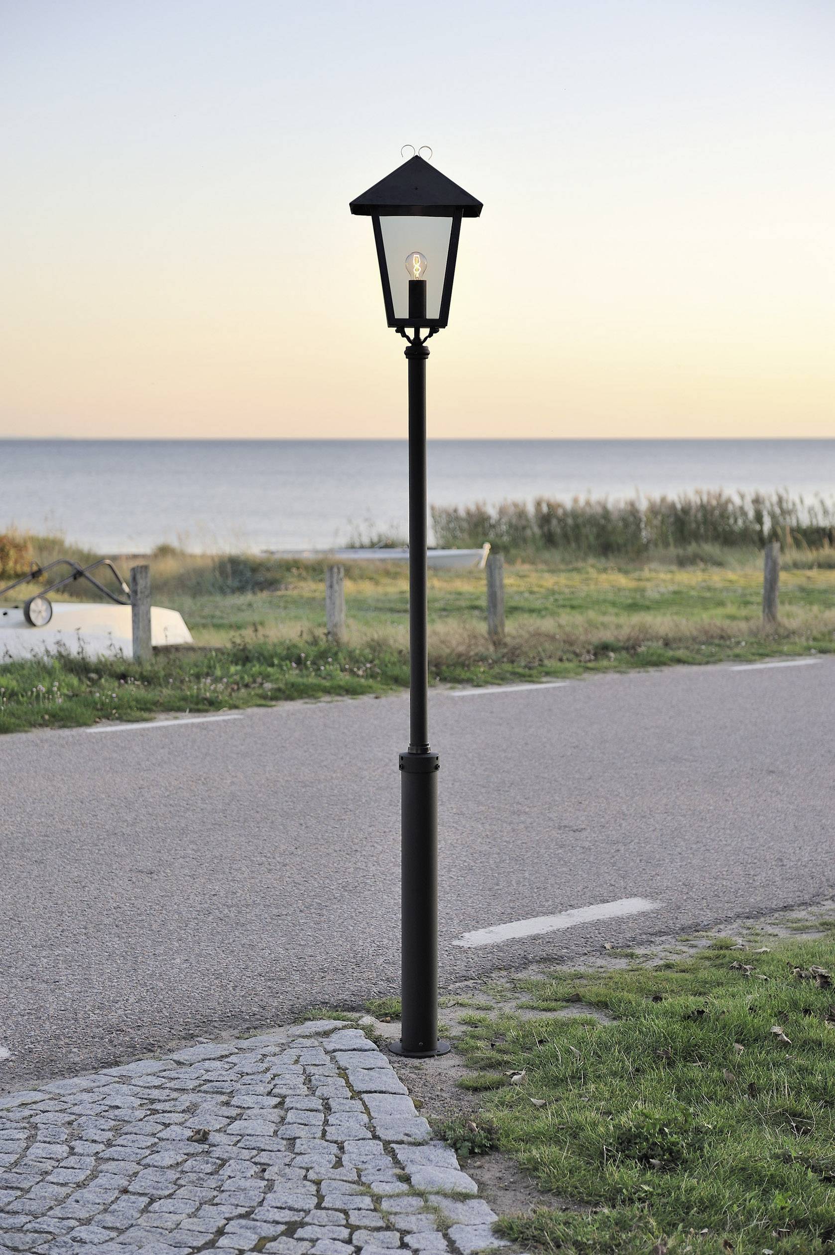 Latarnia uliczna bezpośrednio przy asfaltowej drodze, w tle morze o zachodzie słońca.