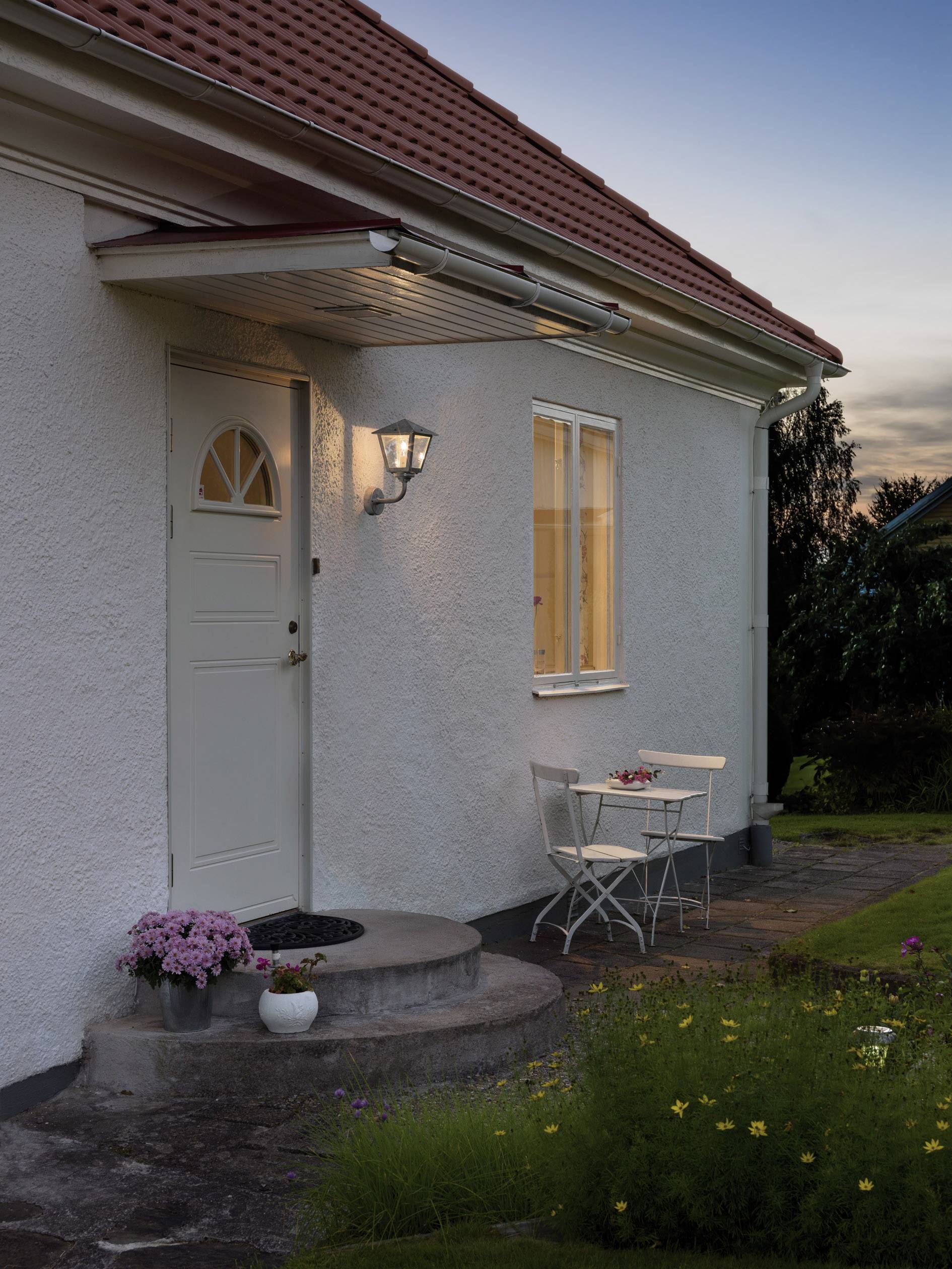 Przytulny dom o zmierzchu, oświetlone wejściowe schody z doniczkami kwiatowymi, obok mały stolik i dwa krzesła.