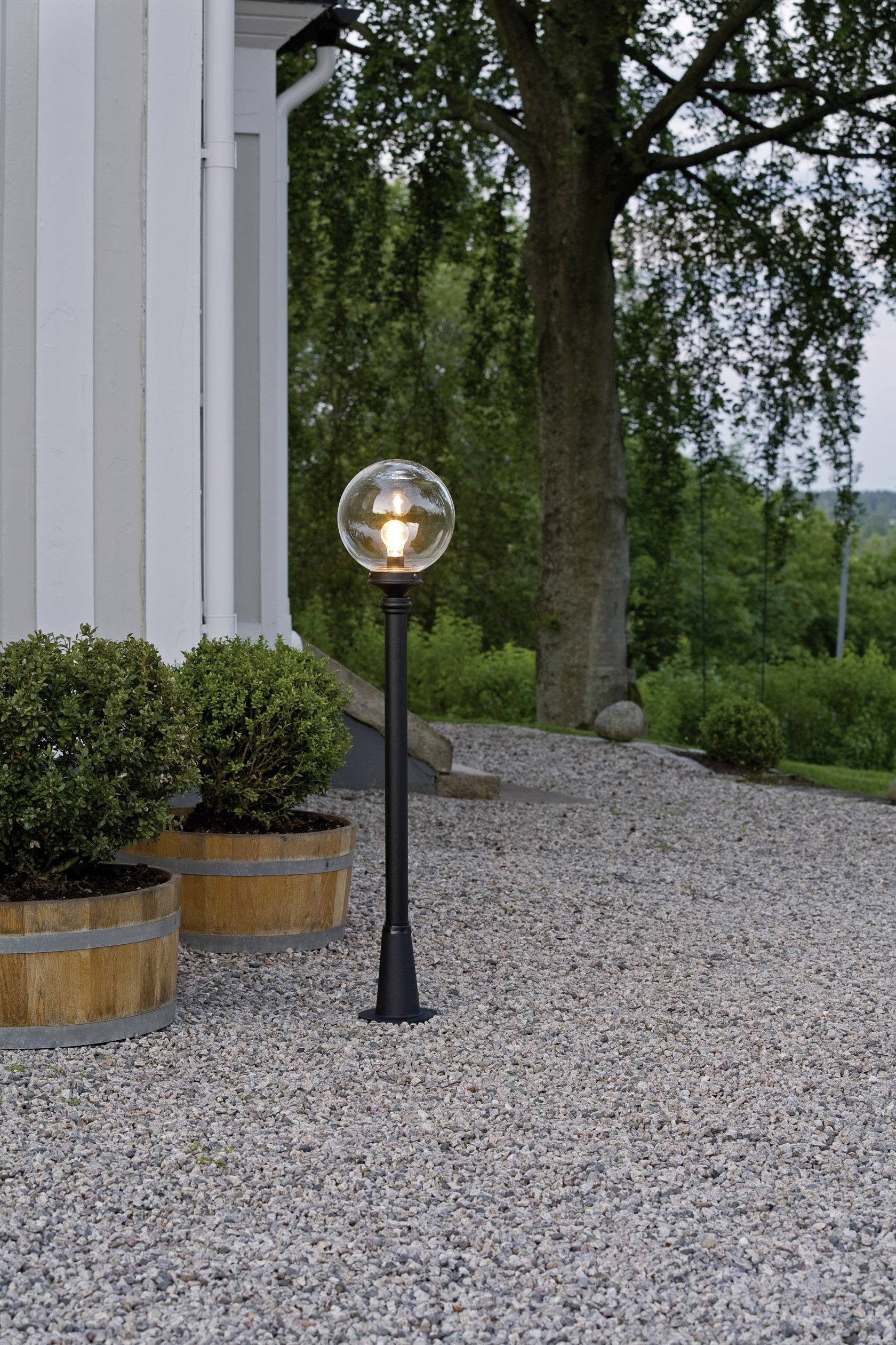 Lampa kulista na żwirowej ścieżce obok białego budynku. Obok stoją rośliny w drewnianych beczkach, w tle widoczne są drzewa.