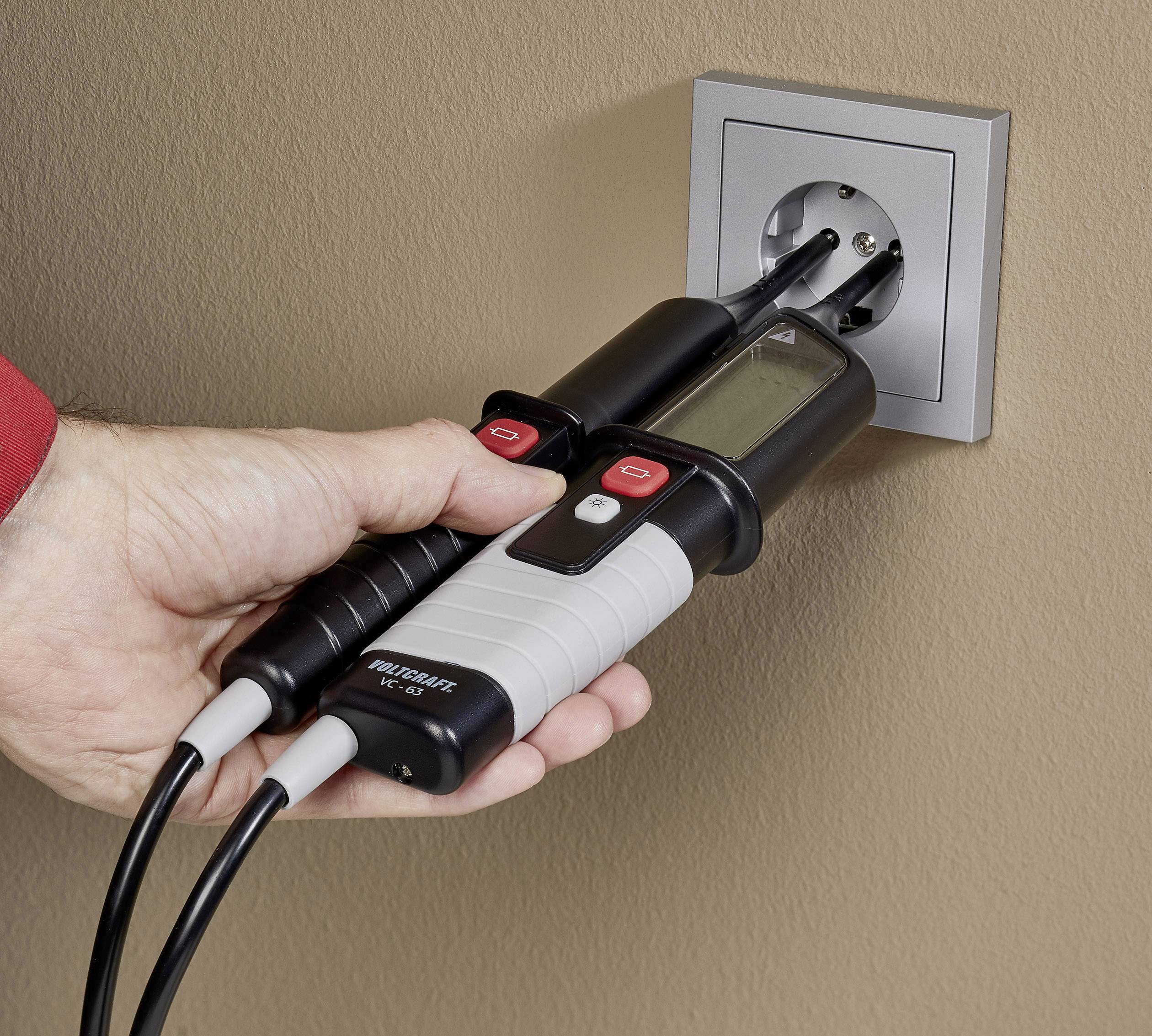 Ręka sprawdza gniazdko elektryczne za pomocą miernika napięcia. Miernik wyświetla cyfrowy odczyt. Ściana jest w kolorze beżowym.